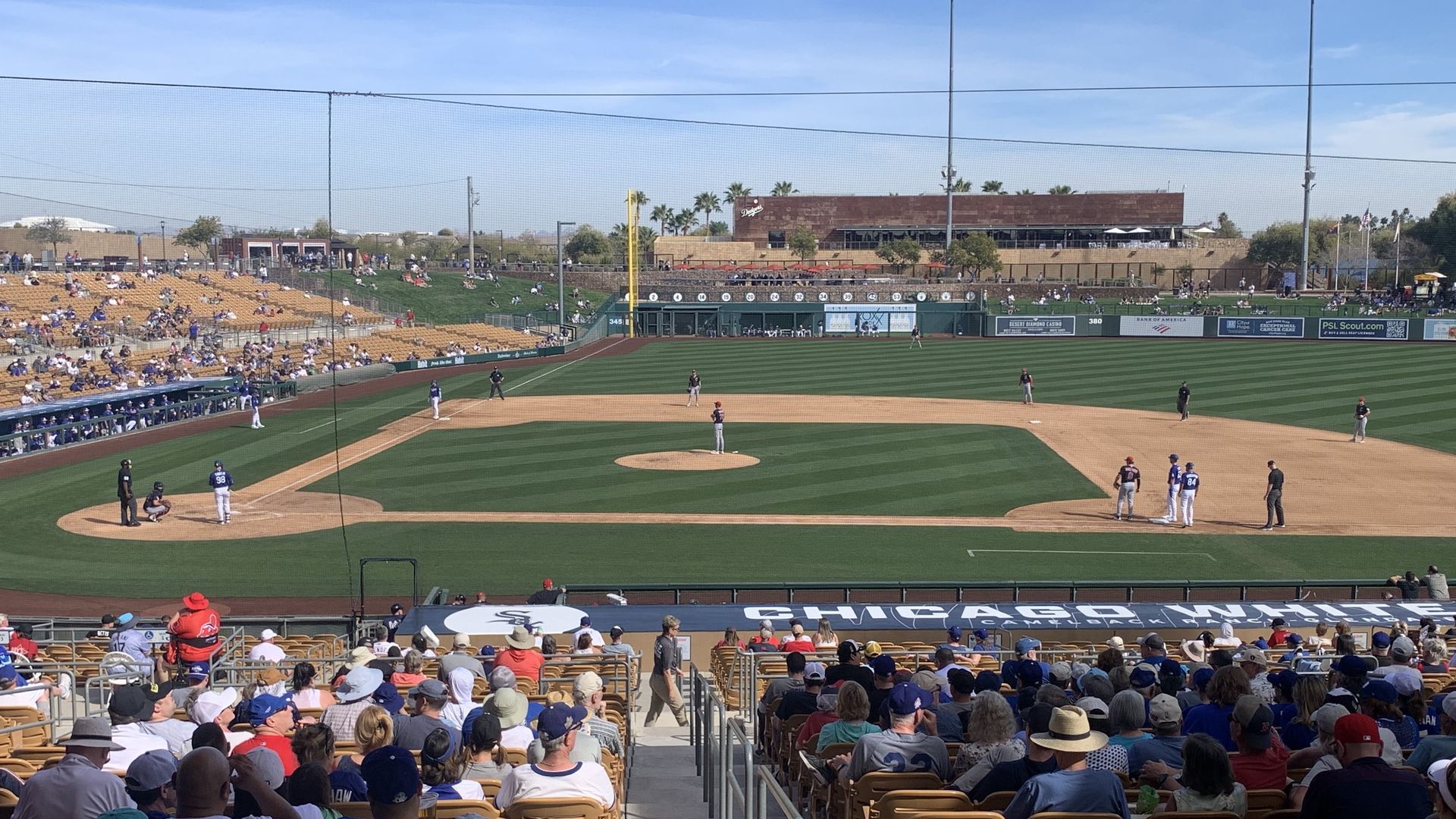 Baseball game at a sunny stadium with players on the field, fans seated in tan chairs, and blue sky. Sign on railing reads "Chicago White Sox."