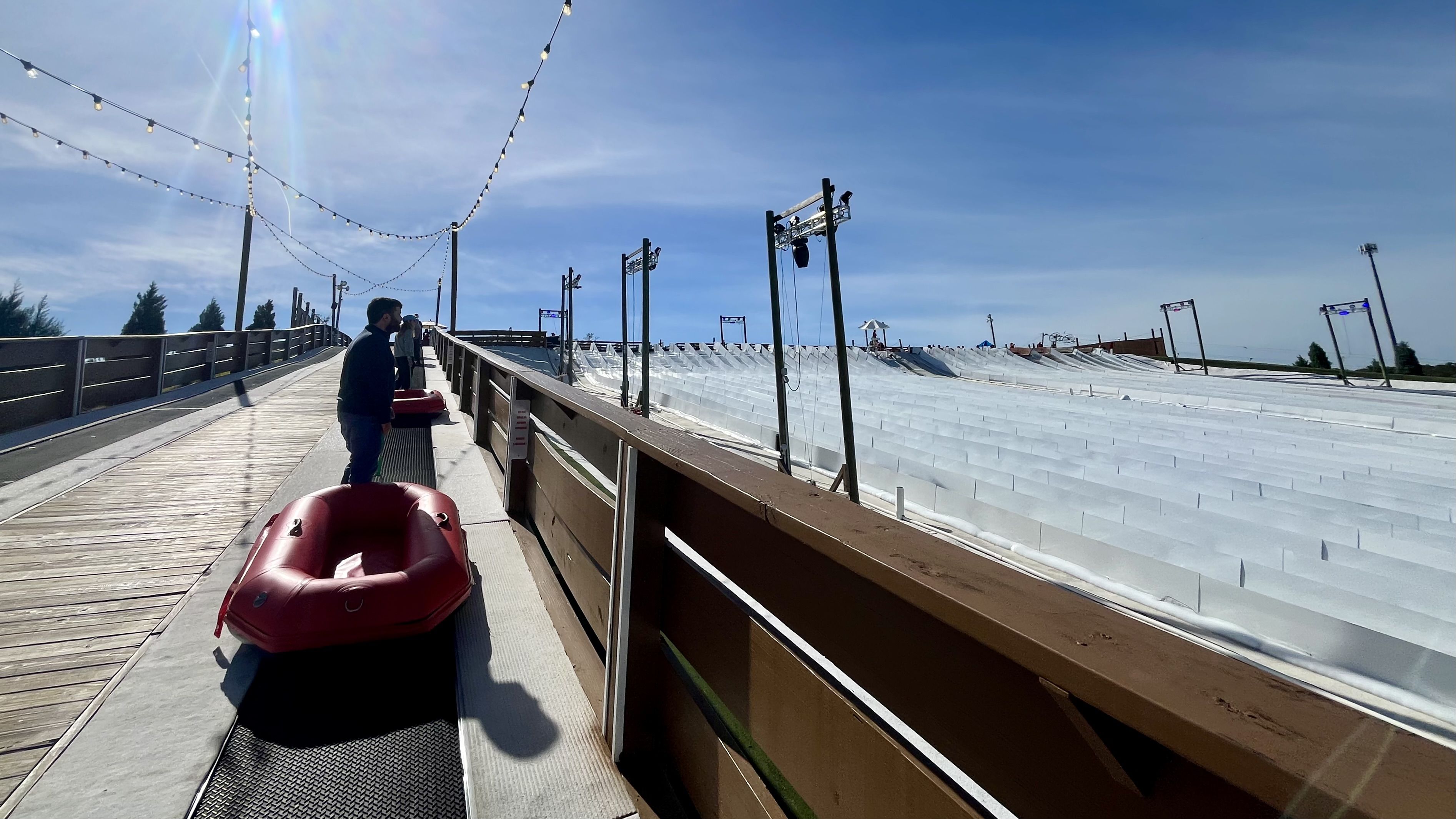 People with red inflatable tubes stand on a conveyor belt ramp next to multiple wide white lanes for sliding under a sunny blue sky with string lights overhead.