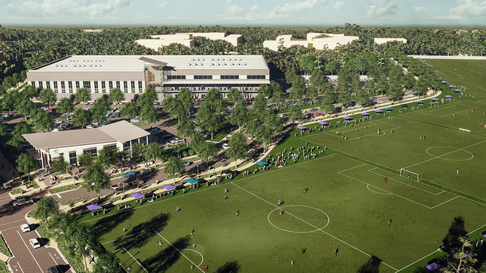 Aerial view of a large green soccer field with players, surrounded by trees and spectators under blue and purple canopies, adjacent to two modern buildings and a parking lot.