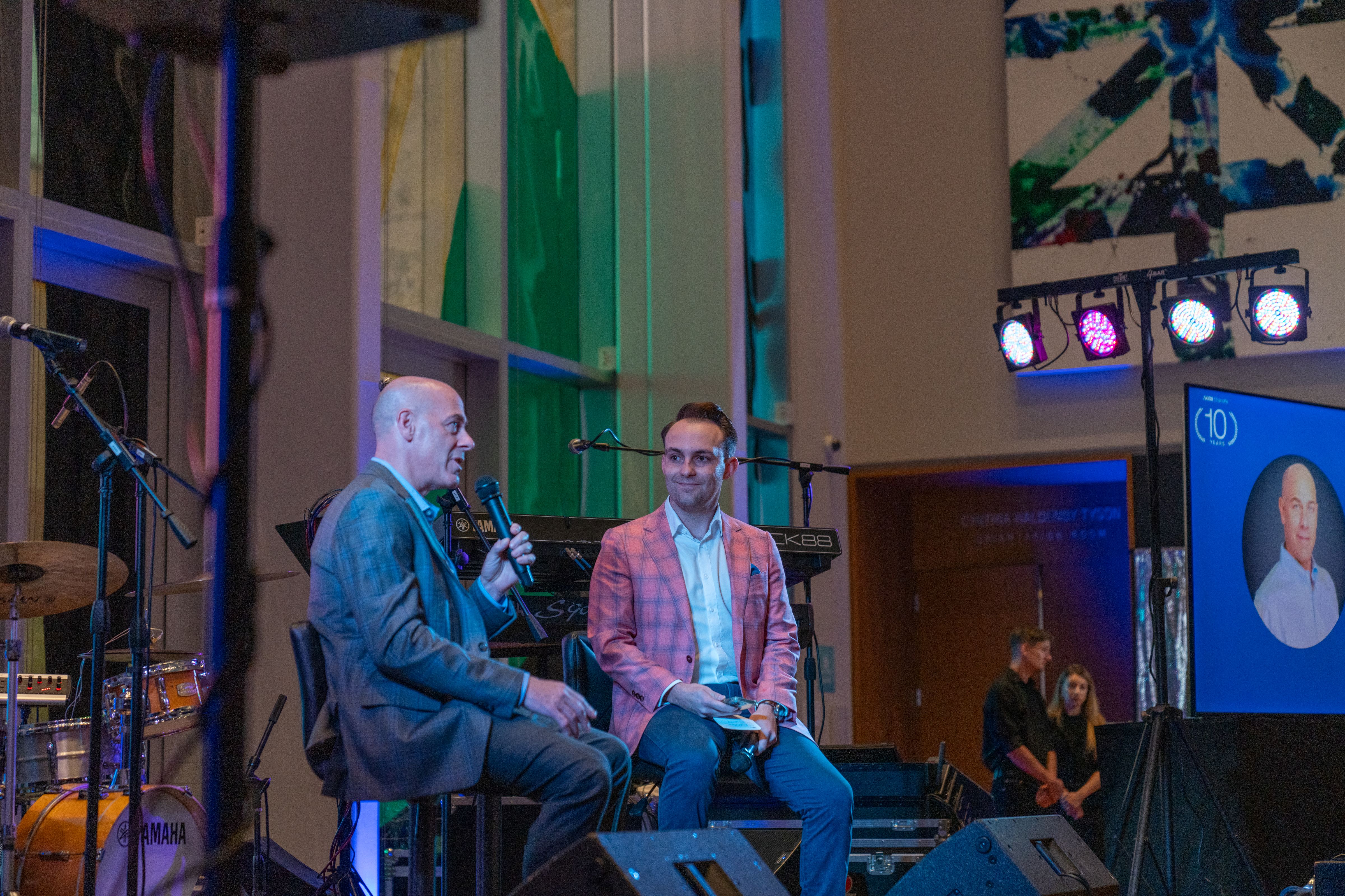Two men in suits sitting on stage with microphones, one in a gray plaid suit speaking, the other in a pink plaid jacket listening. Colorful stage lights and music equipment around.