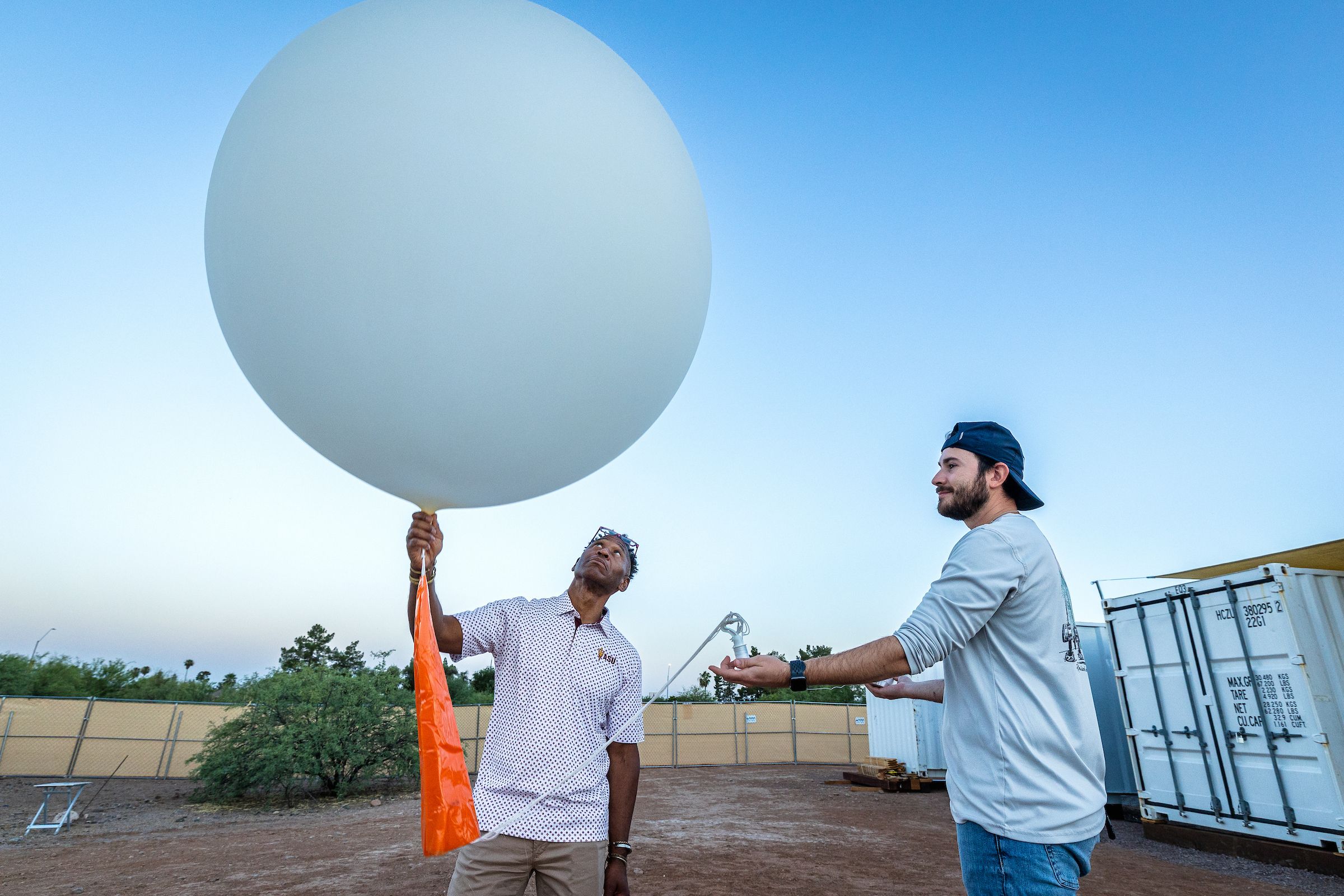 Two men release a large white balloon outdoors under a blue sky. One holds the tether while the other extends a hand; containers and a fence are visible in the background.