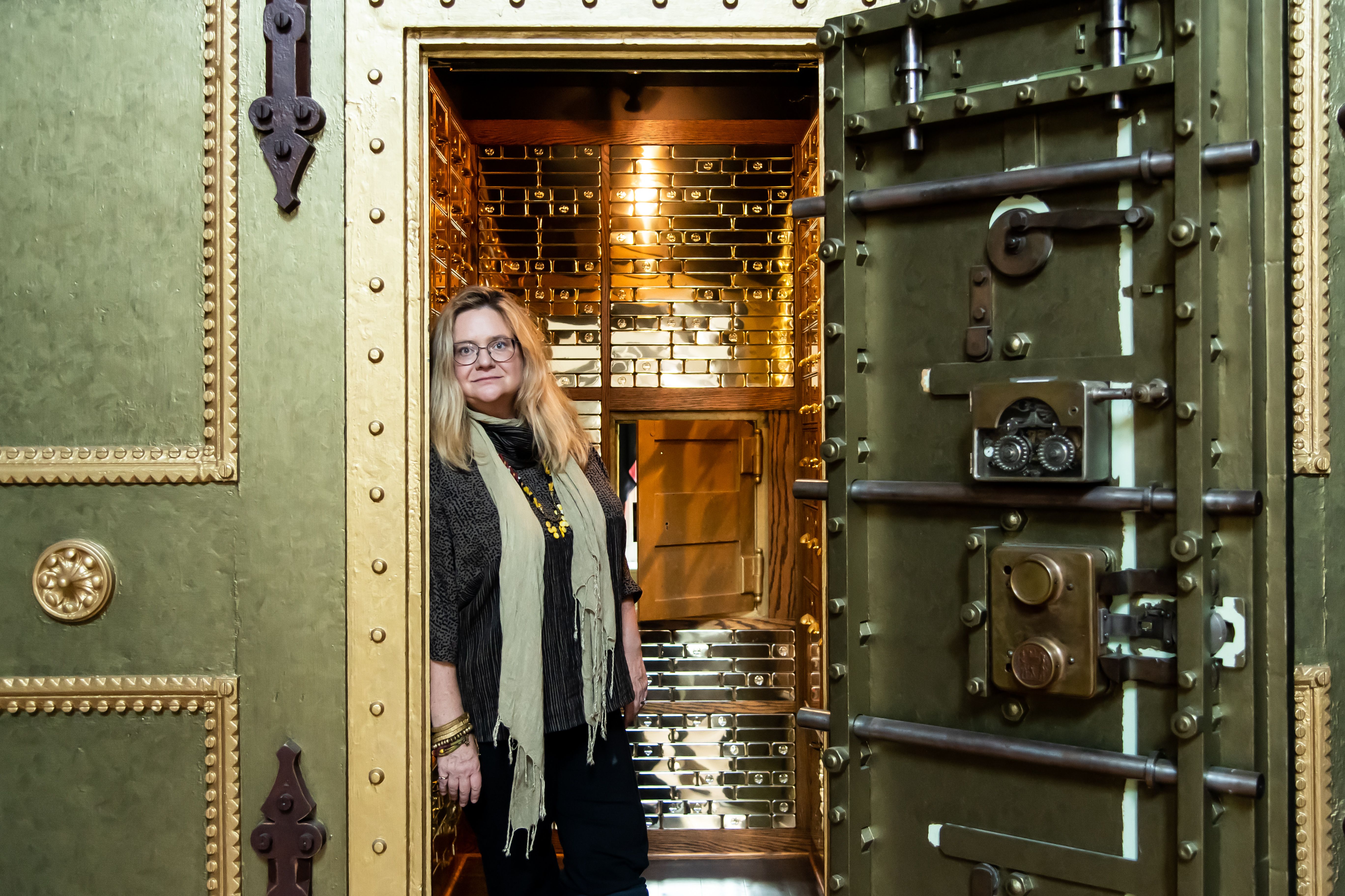 A woman with glasses, blonde hair, and a beige scarf stands inside an open large green vault door, revealing a shiny gold interior filled with metal boxes.