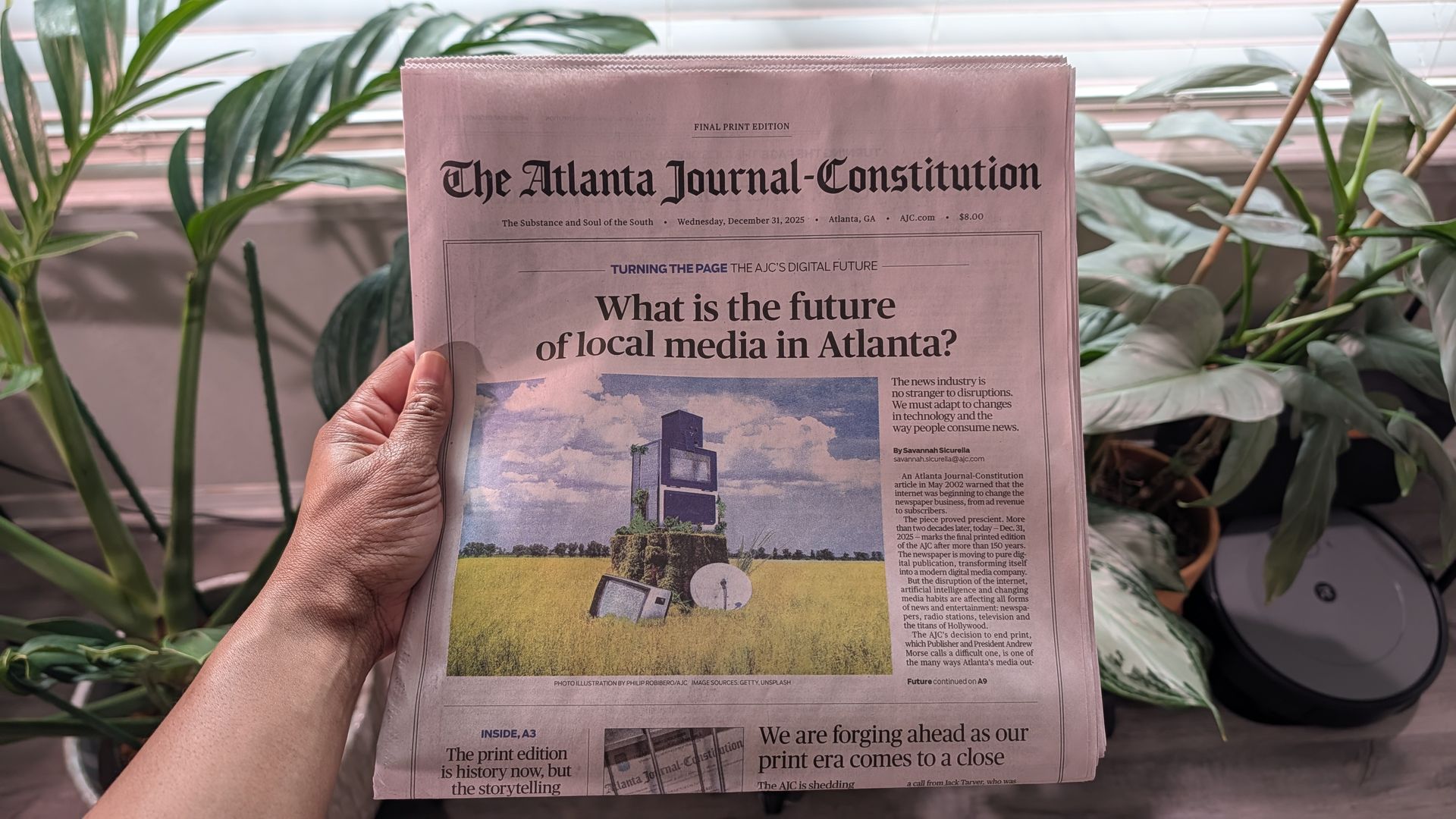A hand holding the final print edition of The Atlanta Journal-Constitution newspaper dated December 31, 2025, with headline about the future of local media in Atlanta, surrounded by green plants.