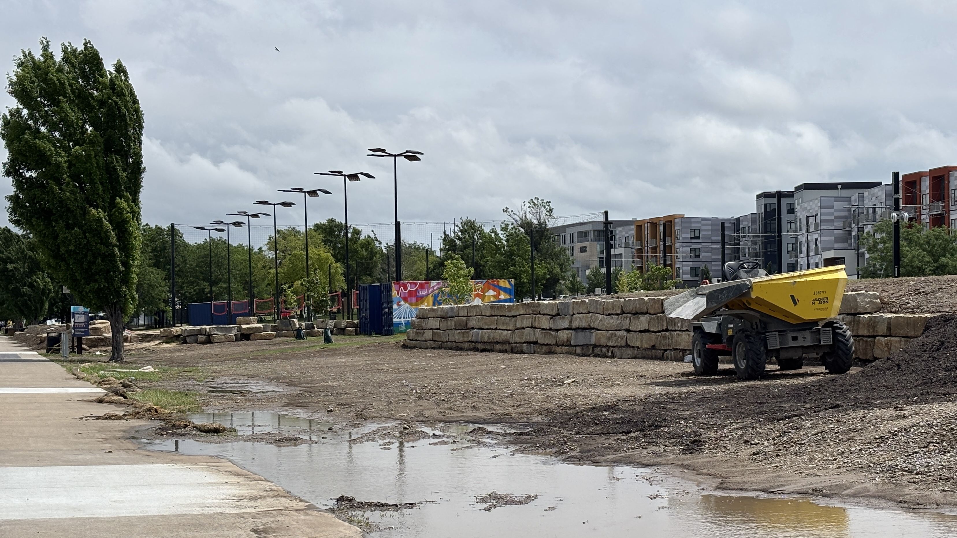 Construction site with a yellow dump truck on the right, a stone wall, and a puddle; modern gray buildings under construction in the background; tall tree left, solar lights along a fence, cloudy sky.