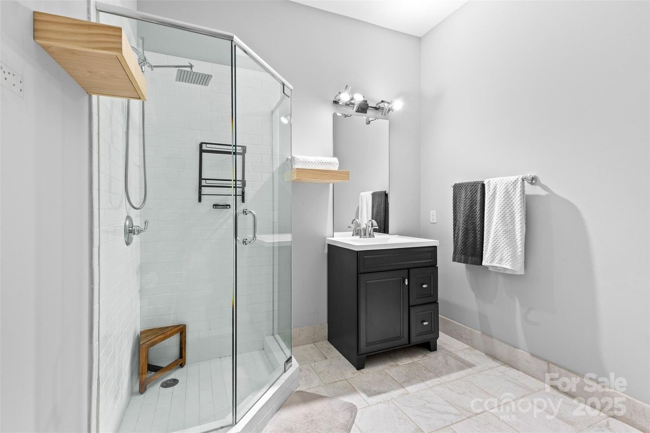 Modern bathroom with glass shower, white tiled walls, wooden corner stool, black vanity with white sink, lit mirror, wooden shelf with towels, and gray and white towels on silver rack.