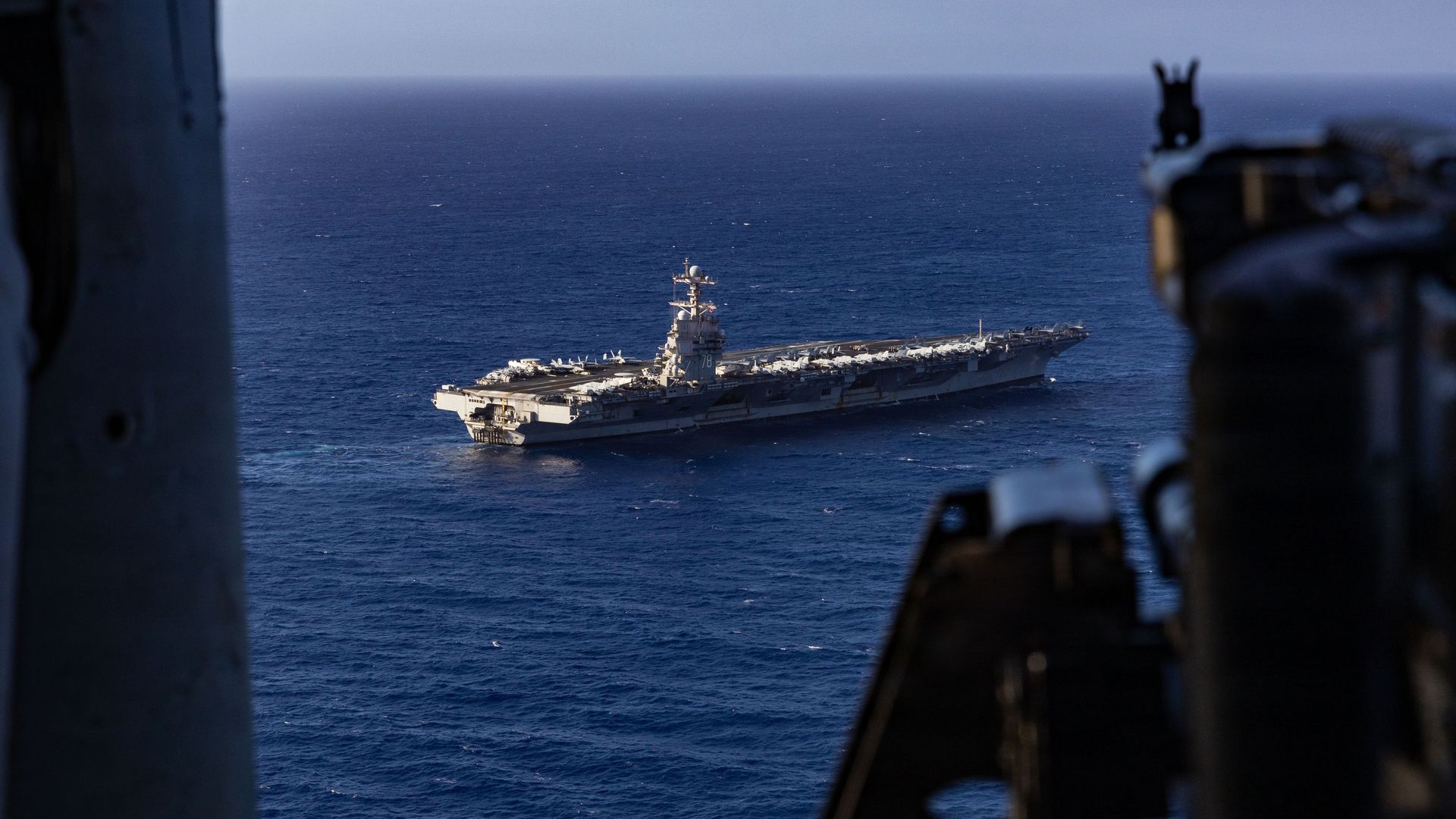 Photo of a large aircraft carrier numbered 78 sailing on a deep blue ocean, seen from behind a dark structure on another vessel.