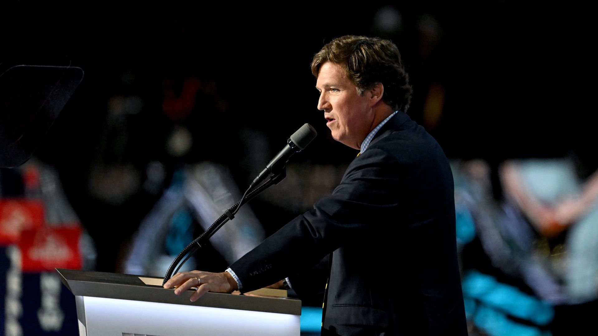 Tucker Carlson, wearing a gray suit and speaking into a microphone at a podium.