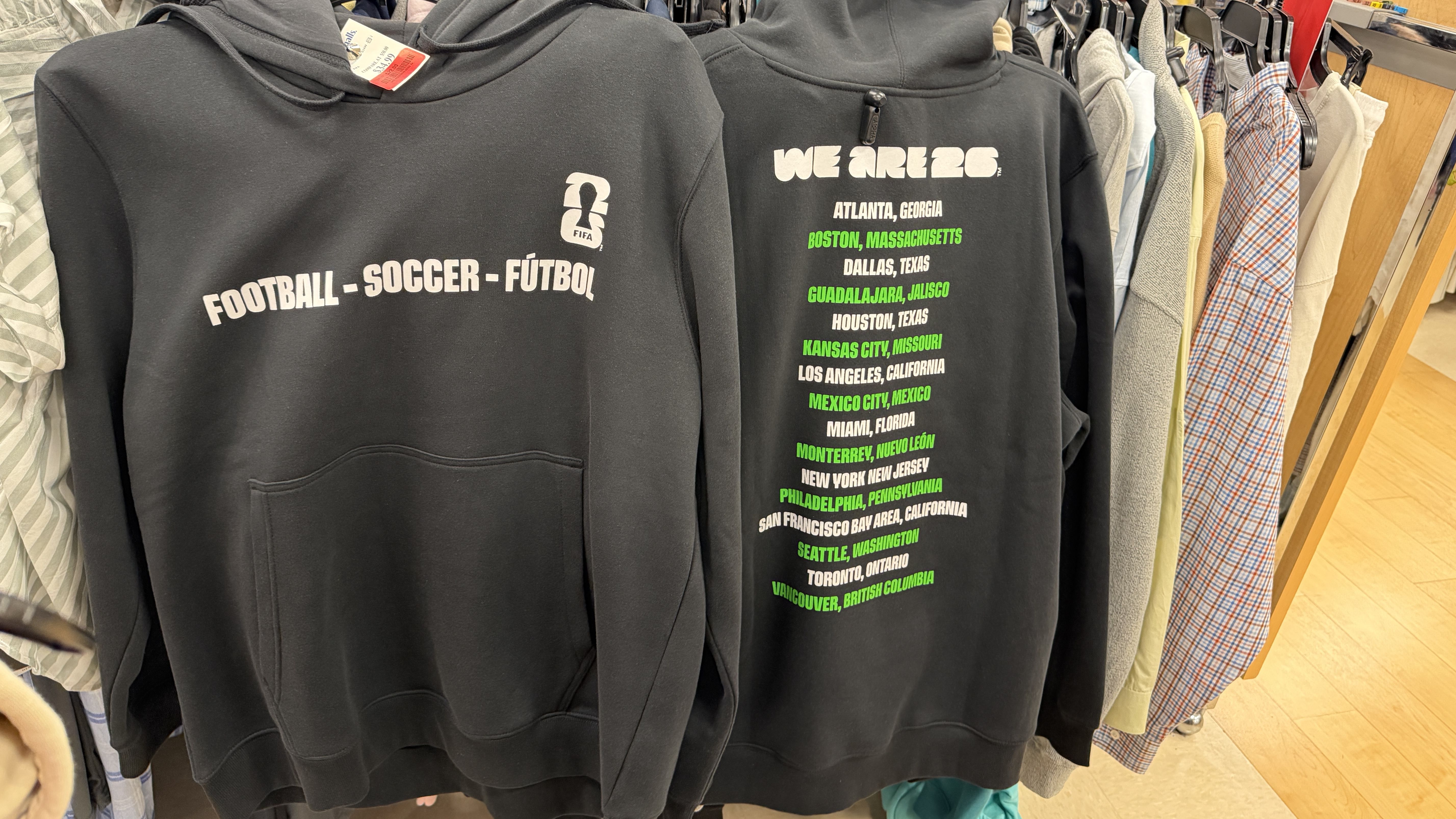 Two black hoodies on a store rack. Left hoodie shows the title "FOOTBALL - SOCCER - FÚTBOL" with a small FIFA logo; right hoodie reads "WE ARE 26" and lists cities in white and bright green.