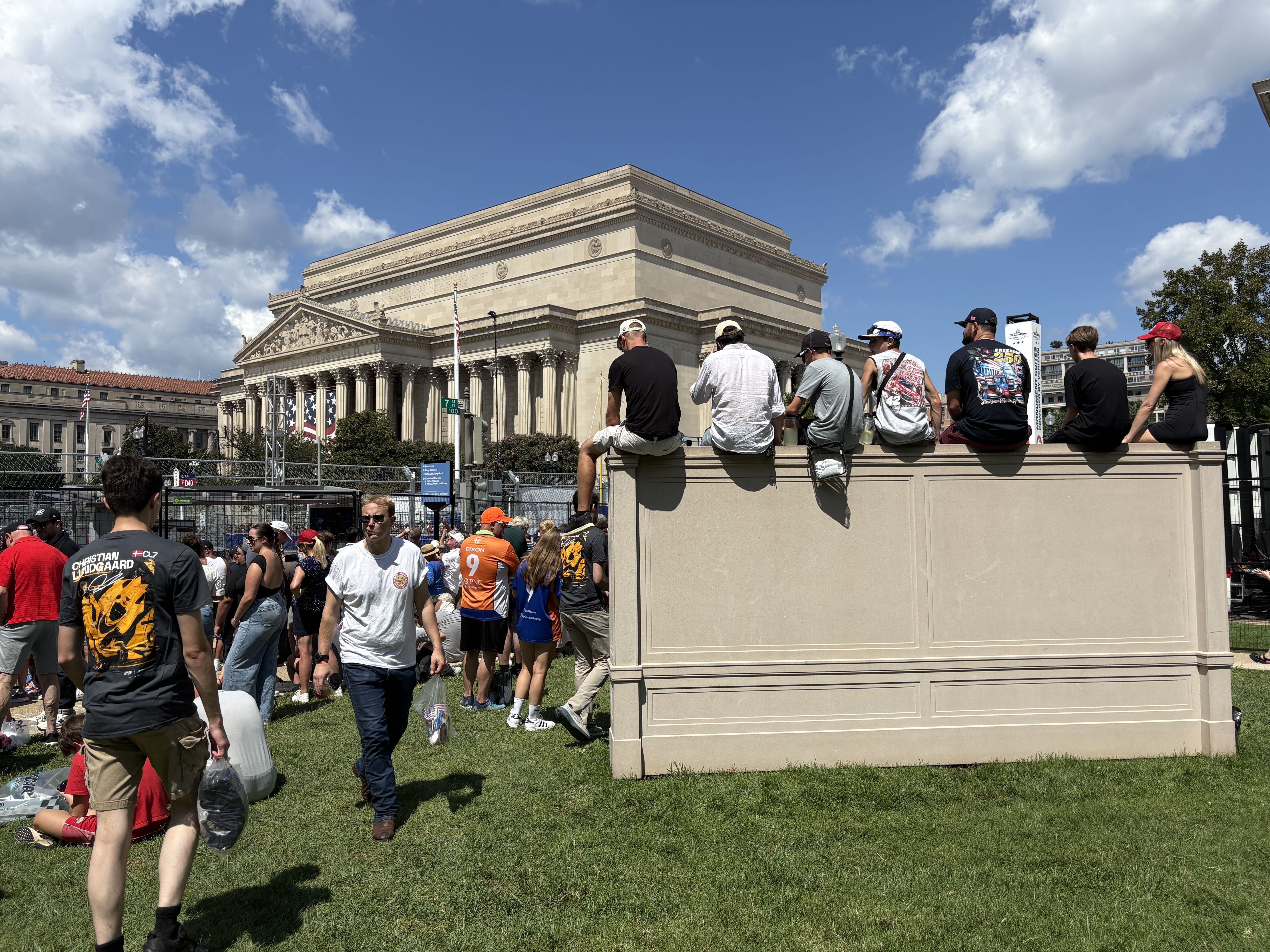 Crowd of people gathered along a beige barrier in front of a large neoclassical building with tall columns; blue sky with scattered white clouds overhead.