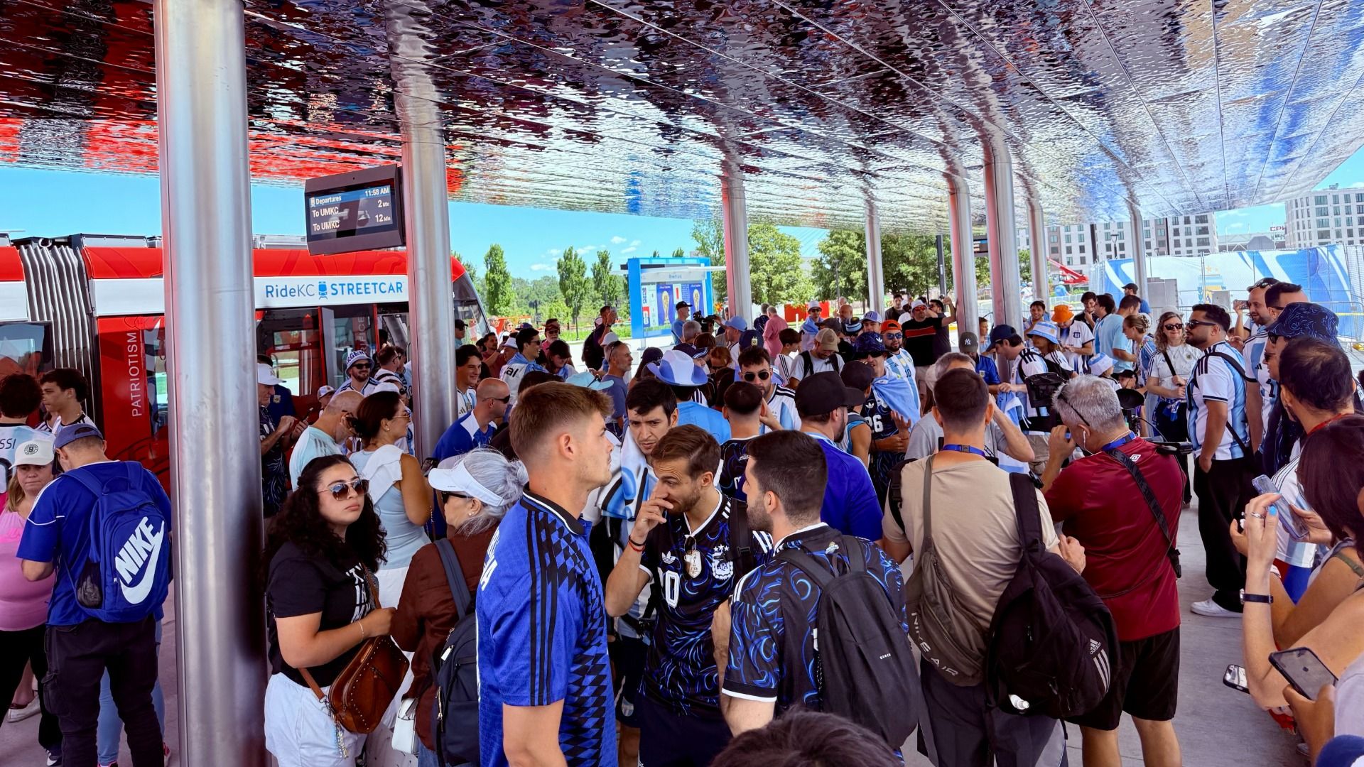 Crowded outdoor station under a reflective ceiling with silver pillars; fans in blue jerseys gather around a red RideKC Streetcar, with a city skyline visible in the background.