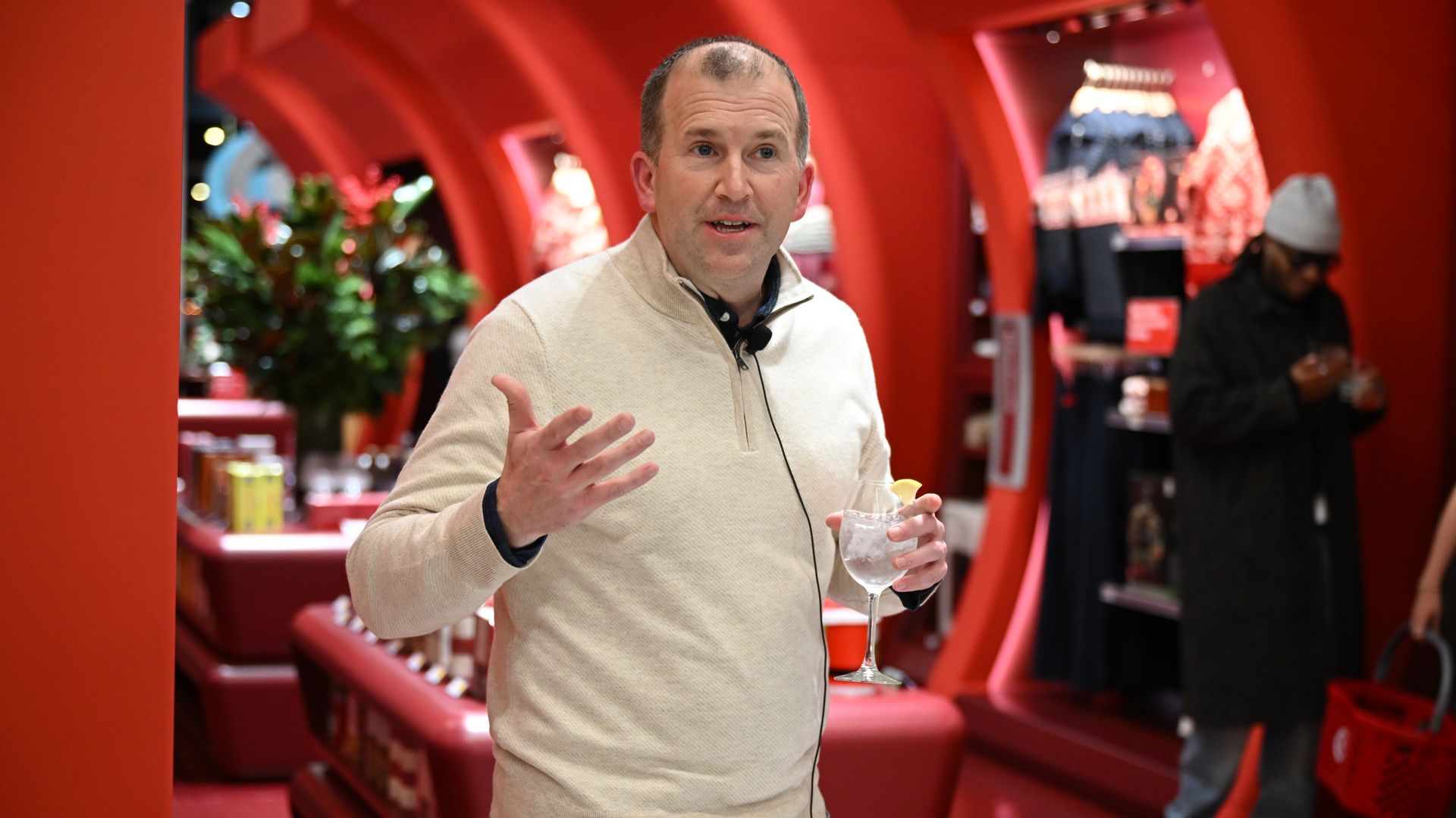 Person in beige sweater holding a glass with ice and lemon, speaking inside a red-themed store with modern curved displays and another person shopping in the background.