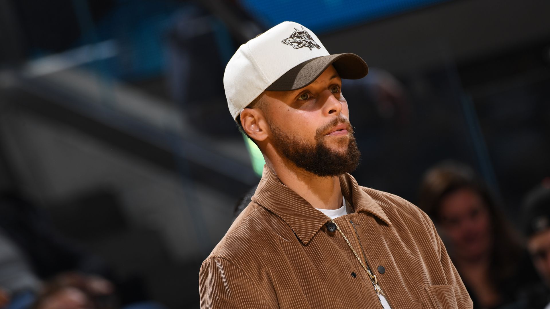 Man with a beard wearing a brown corduroy jacket and white and black baseball cap with a wolf logo, looking to the side in an indoor setting with blurred people in the background.