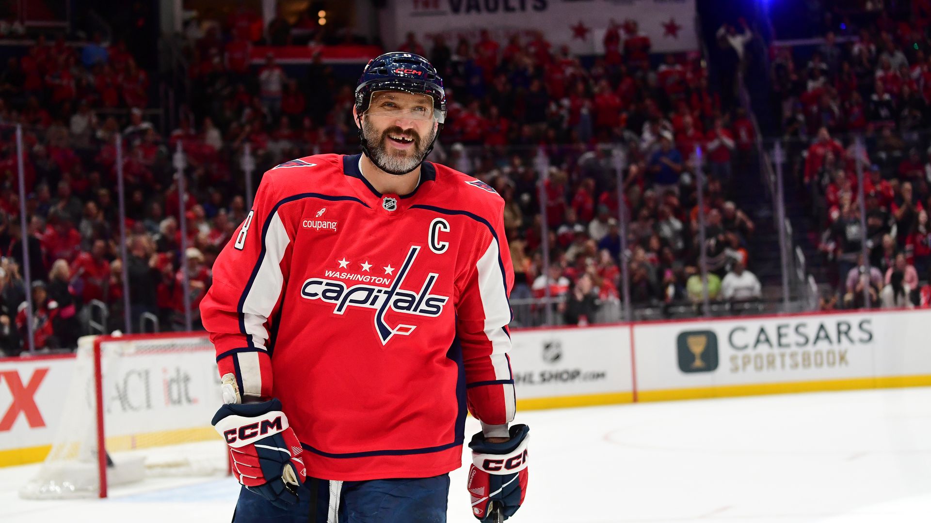 Washington Capitals captain Alex Ovechkin in a red jersey with a C, helmet, and gloves stands on the ice, smiling. A crowded arena in red and a GOAL banner are visible behind him.