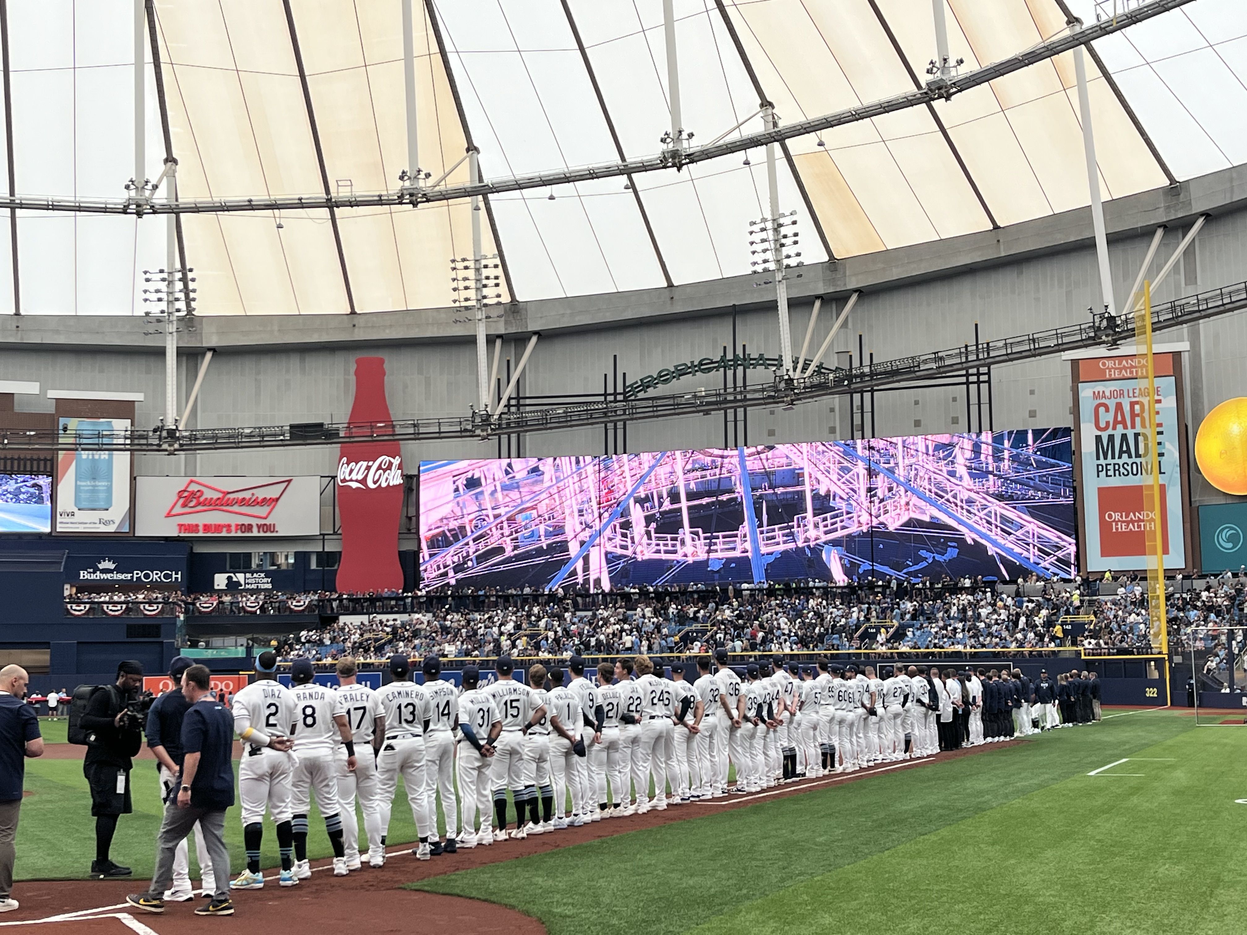 Baseball players in white uniforms stand in a long line along the foul line inside a large domed stadium; a wide LED screen behind them shows a tattered domed roof.
