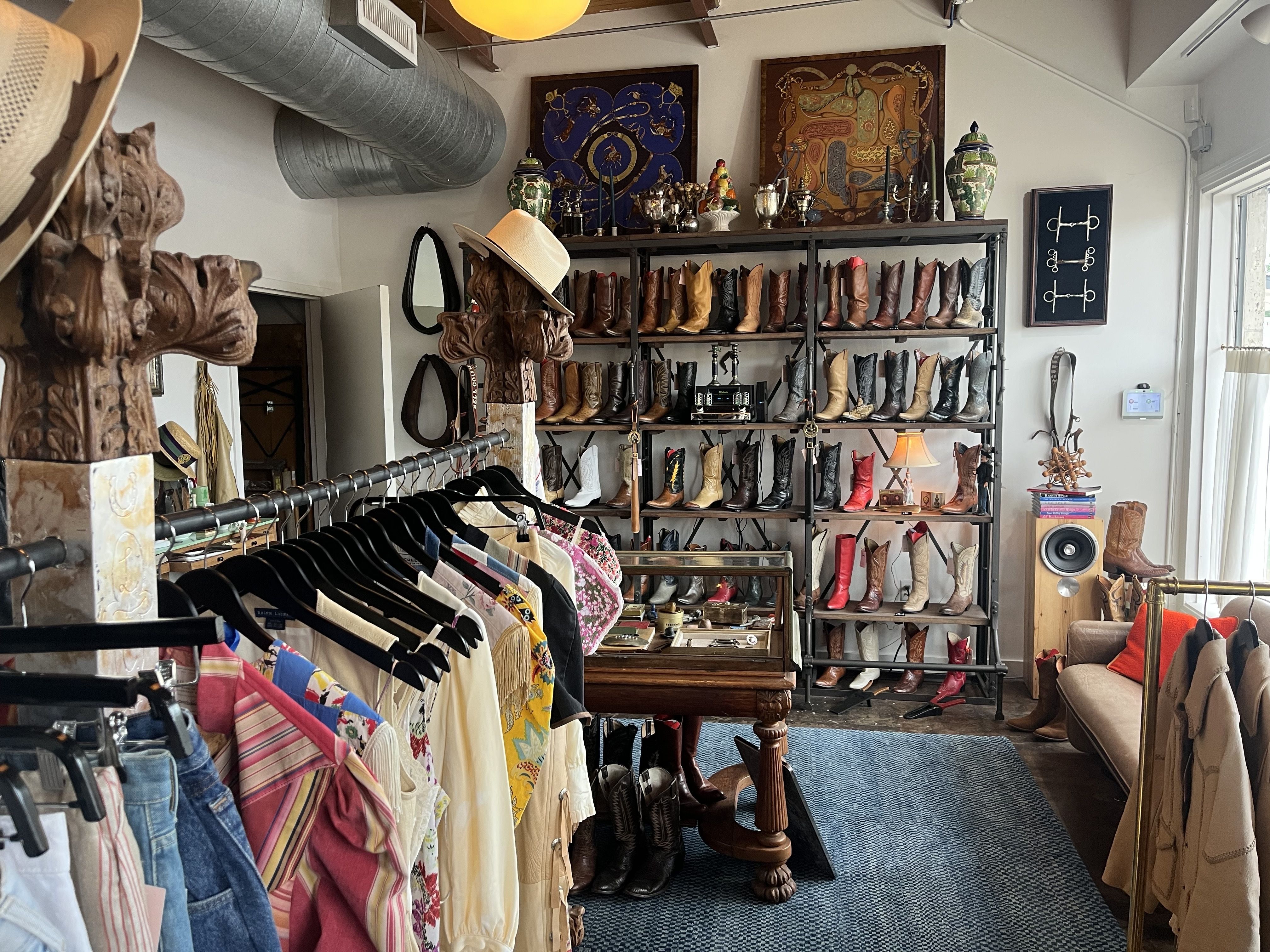 Interior of a western boot shop: shelves of brown, tan, and black cowboy boots; hats, carved statues, framed art, and a clothing rack in the foreground.