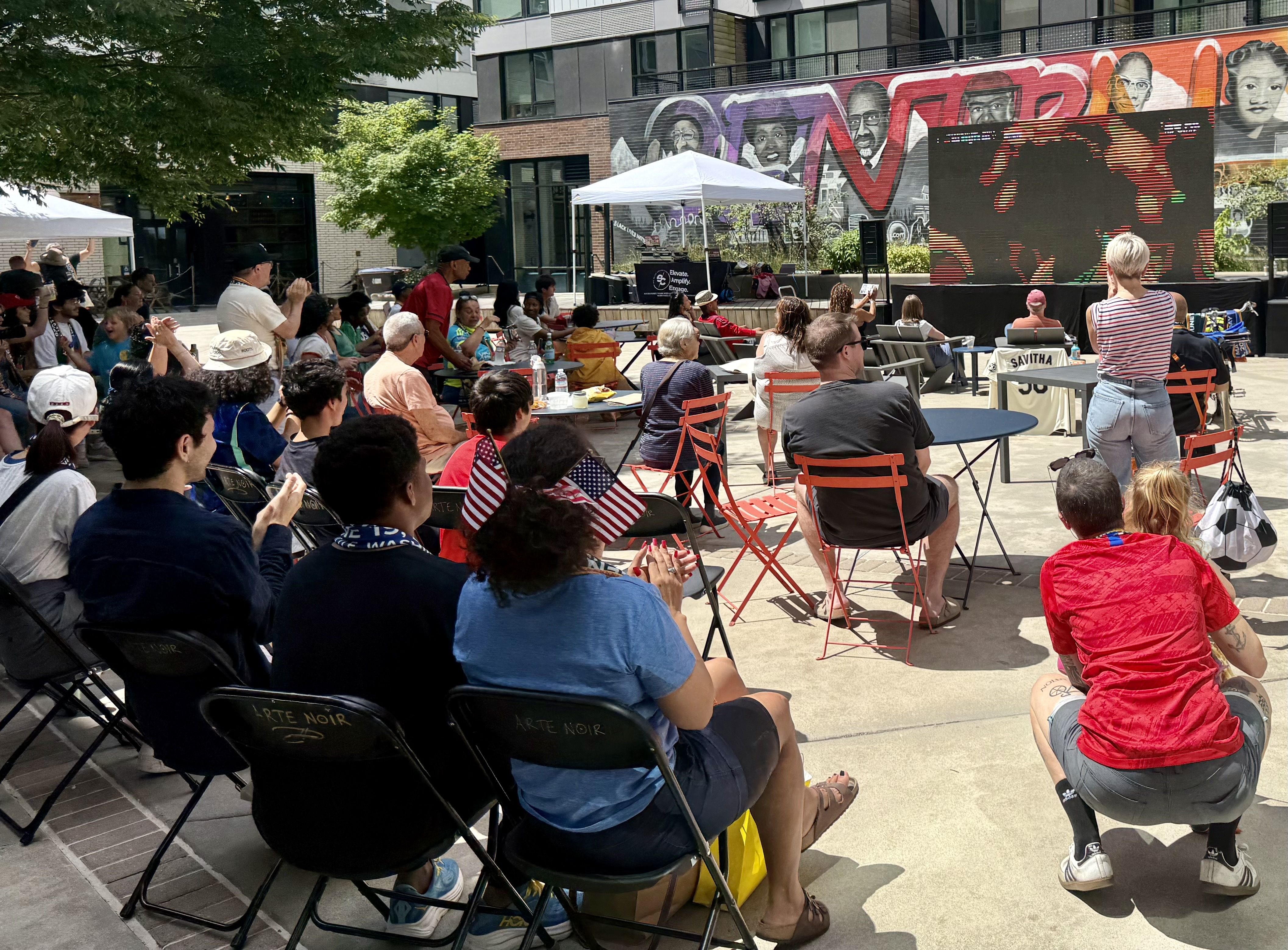 Outdoor community event with a diverse audience seated on folding chairs under white tents. A stage with a large screen shows red patterns, while a mural of portraits decorates the brick wall behind.