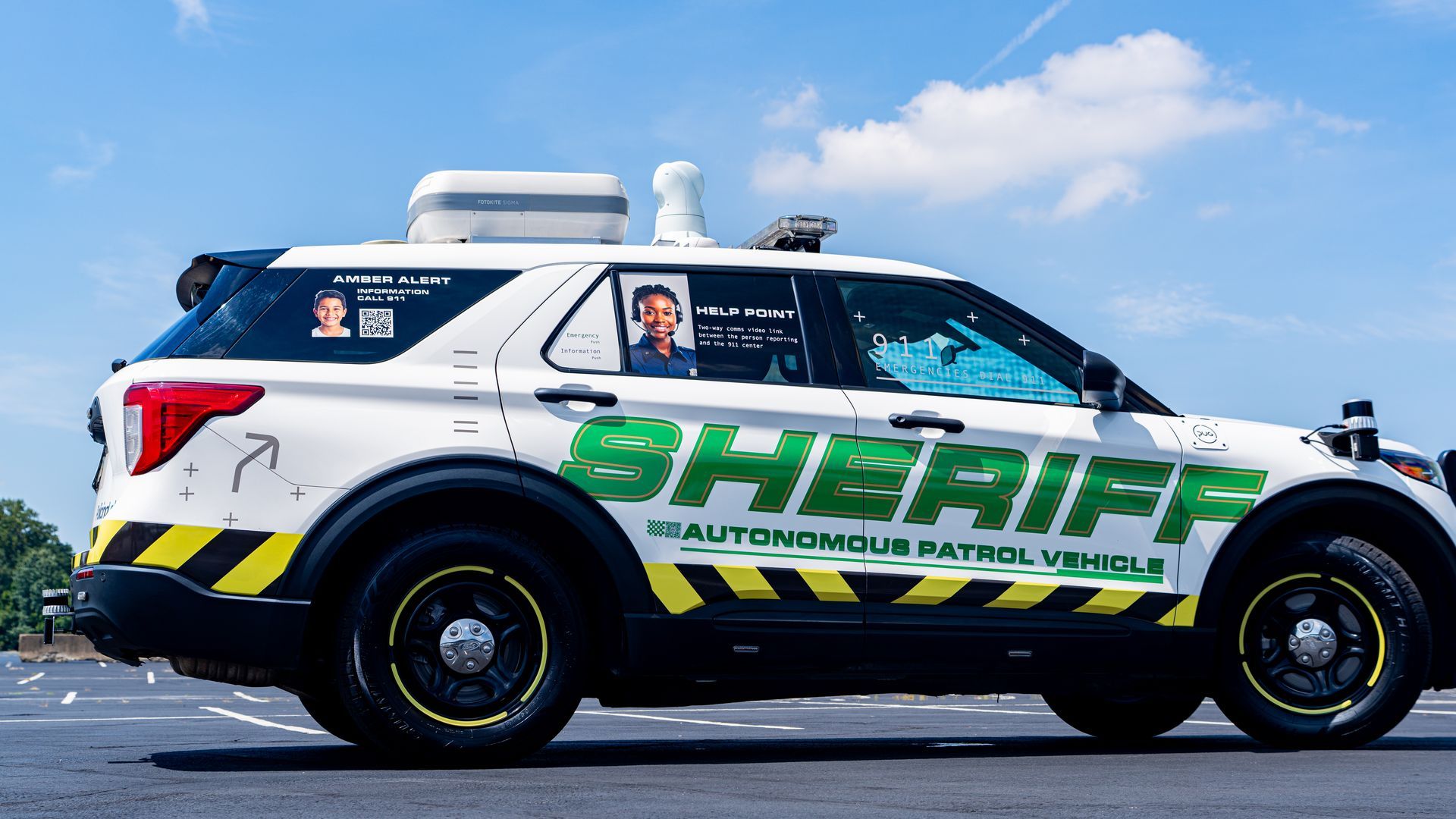 White sheriff autonomous patrol vehicle with green and yellow markings, amber alert photo, and help point video link signage parked outdoors under a blue sky with clouds.