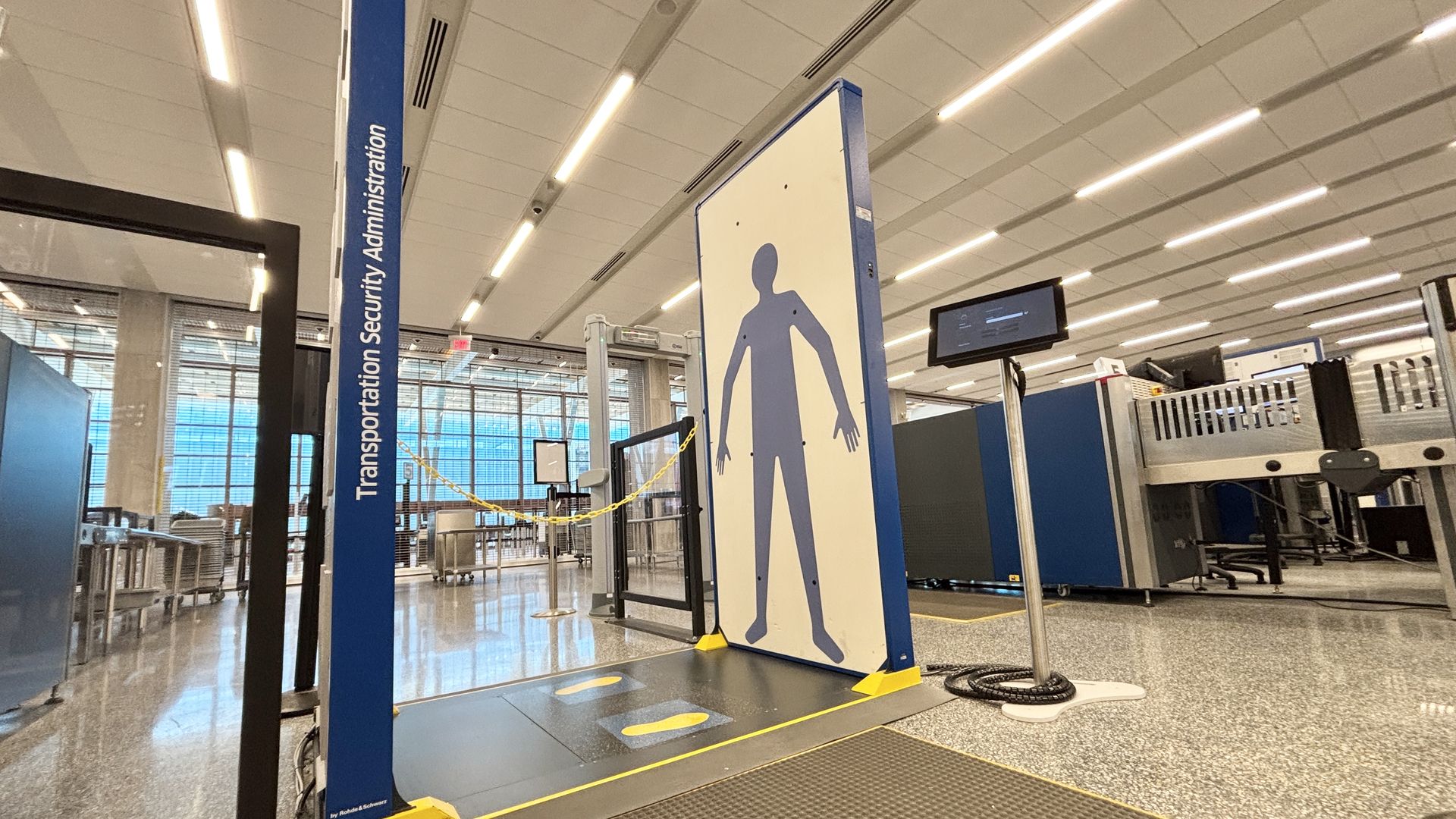 A tall blue pillar labeled Transportation Security Administration stands beside a large panel with a blue silhouette, which are the new imaging technology panels at KCI. The scene includes conveyor belts, barriers, and a monitor under bright lights.
