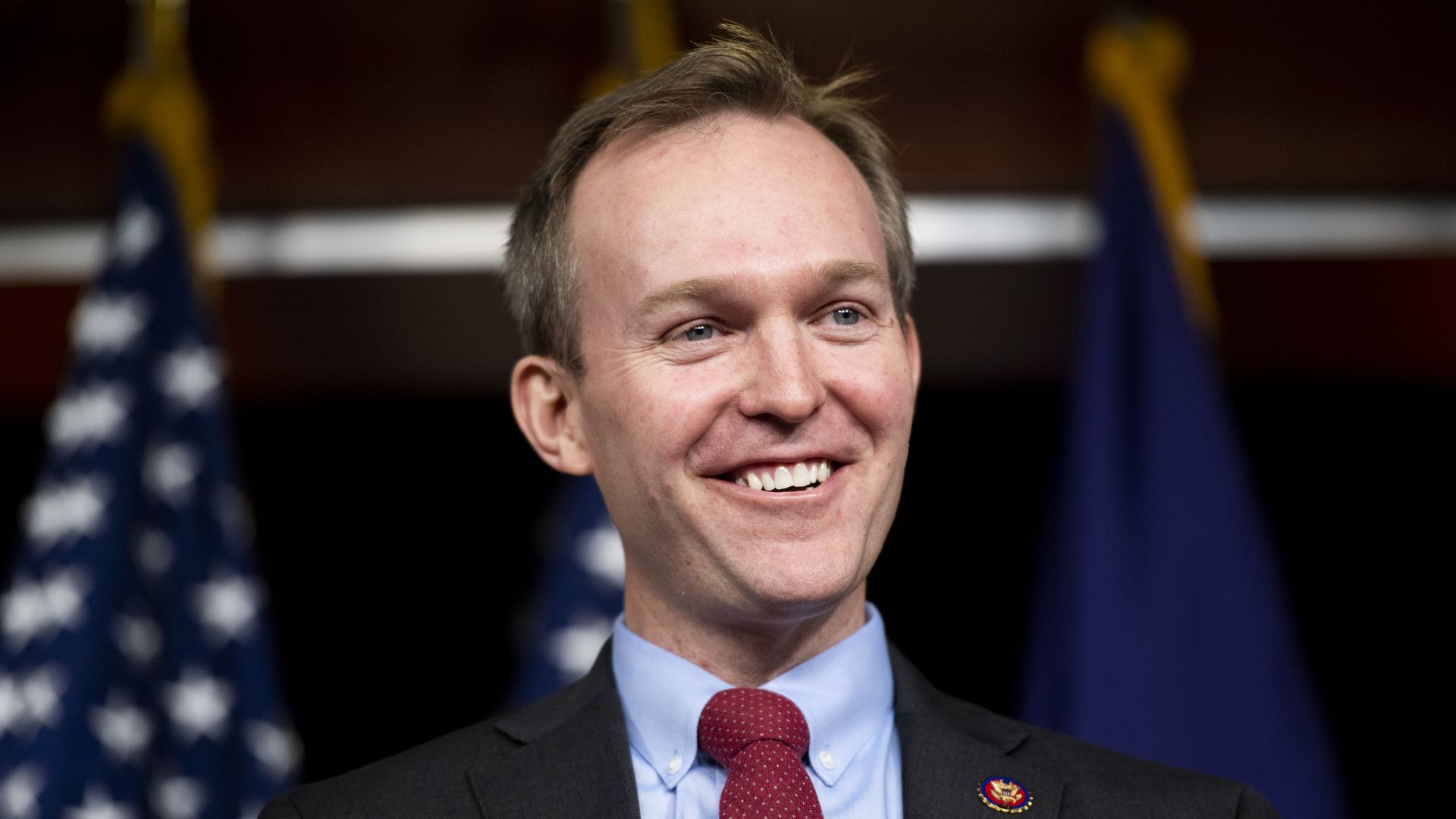 A blonde man in a dark suit and red tie smiles in front of a U.S. flag.