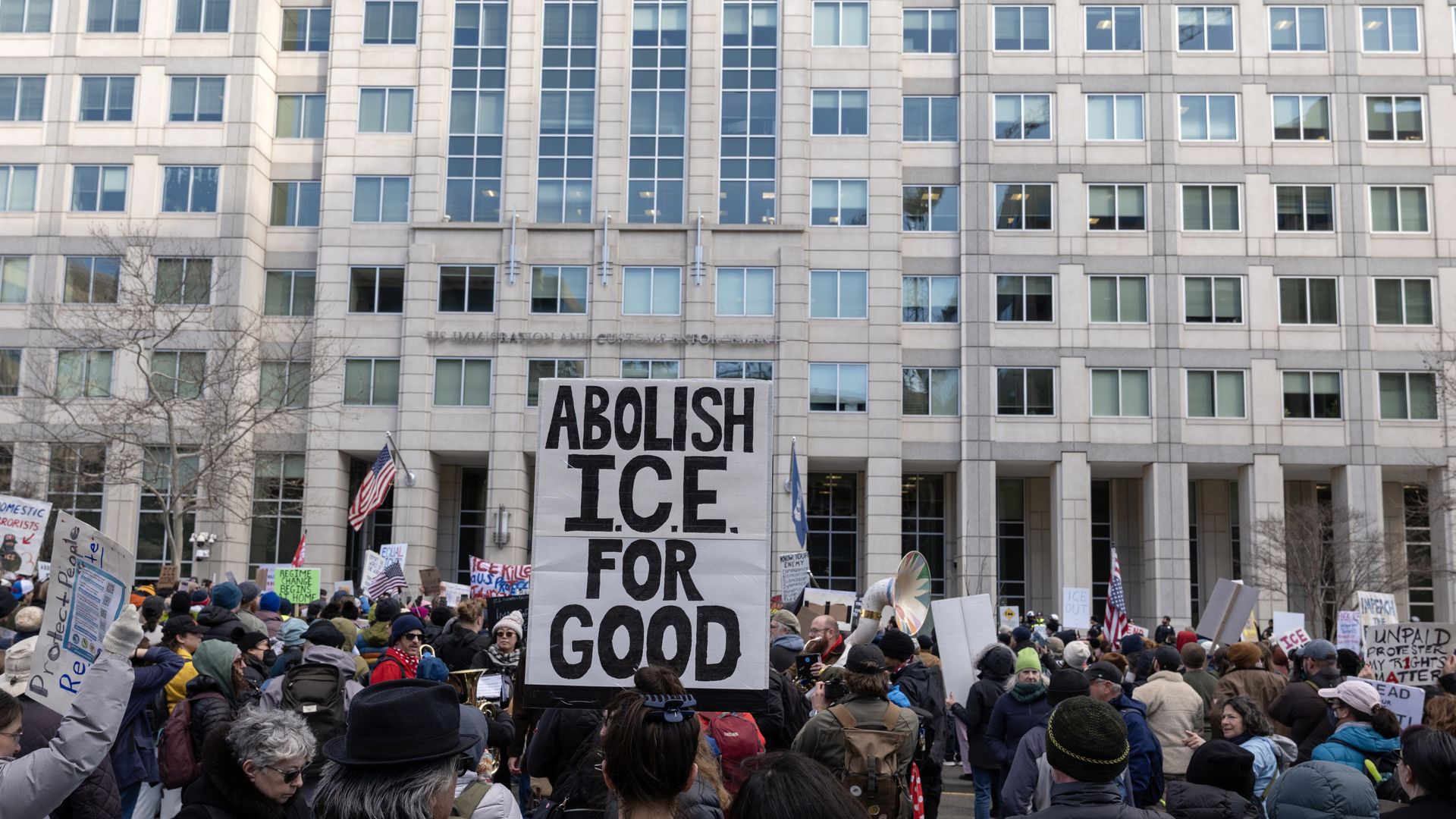 A group of protesters huddles outside of a building as one person holds a sign in the air that reads "Abolish ICE for good"