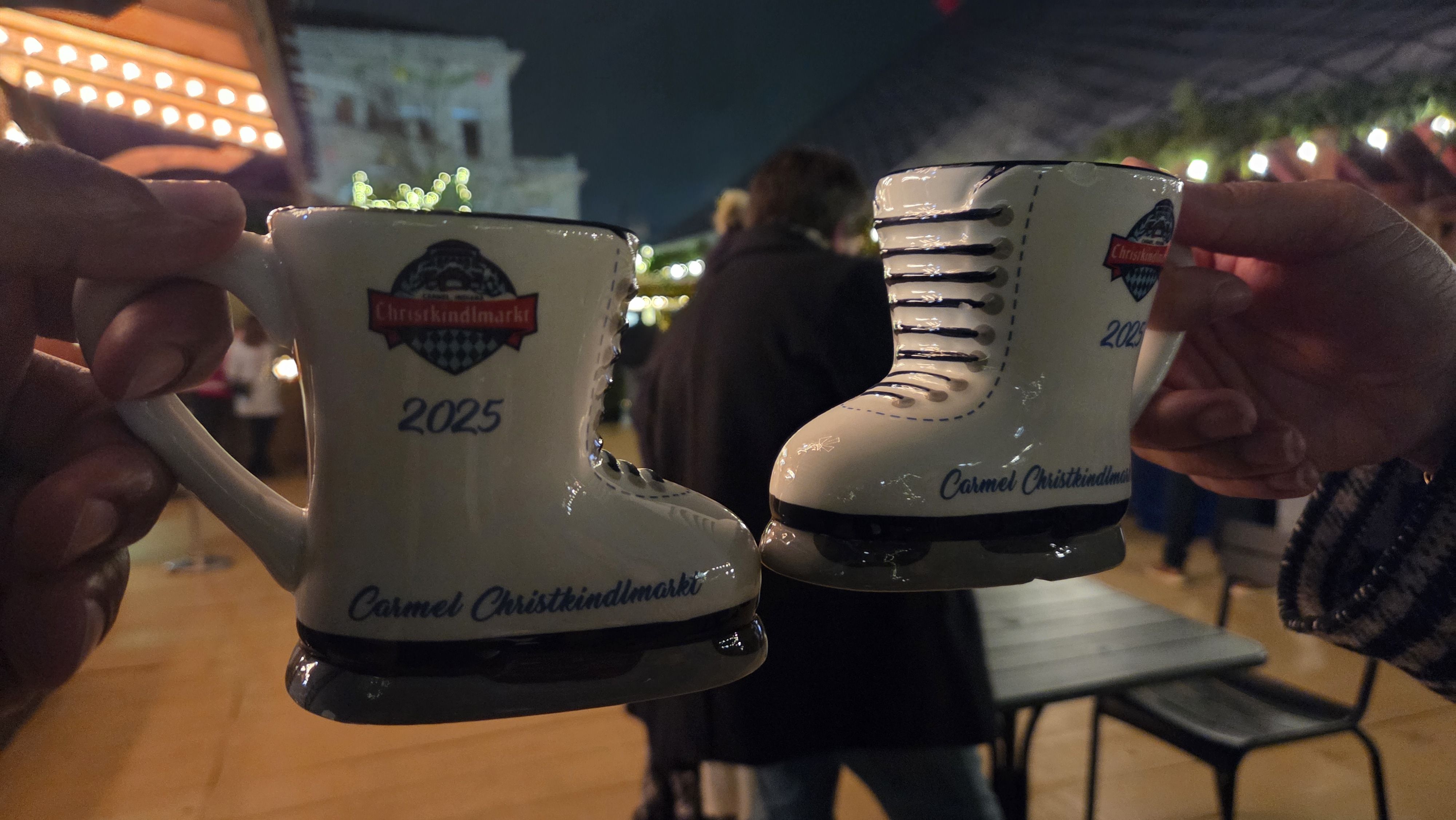 Two hands holding ceramic ice skate cups with "Christkindlmarkt 2025 Carmel Christkindlmarkt" text at a night market with blurred people and festive lights in the background.