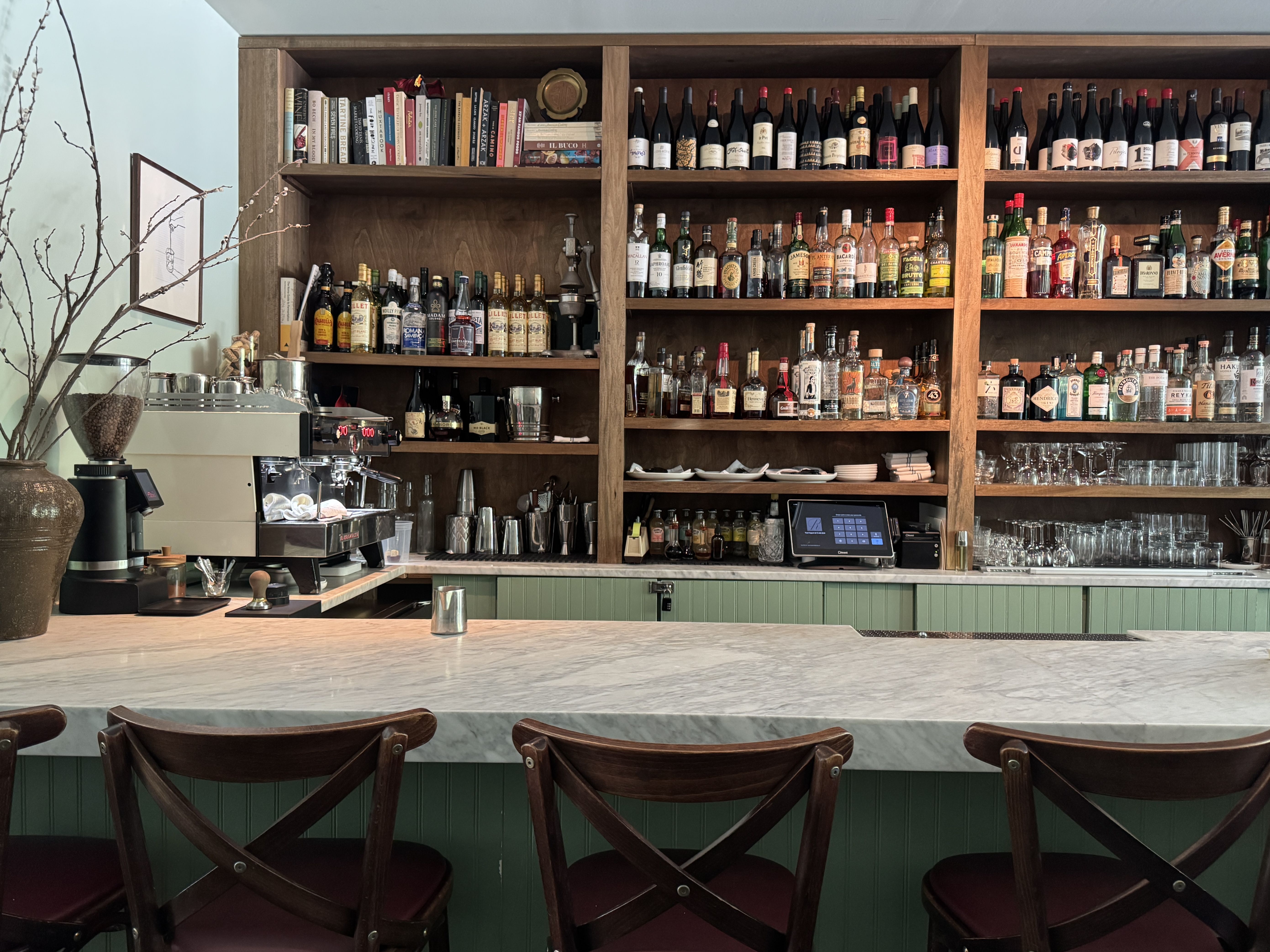 Empty bar counter with three wooden chairs, green paneling, white marble top, shelves filled with wine bottles, spirits, books, glasses, and a coffee machine on the left side.