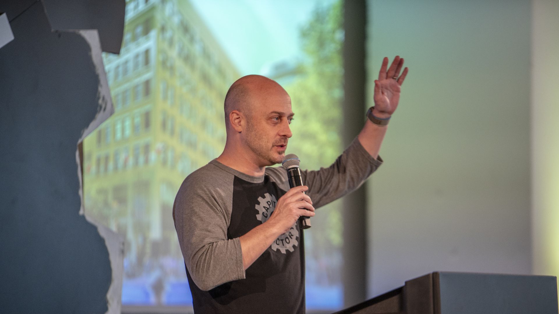 Speaker with a shaved head wearing a gray and black shirt, holding a microphone and raising a hand on a stage; a cityscape projection is blurred in the background.