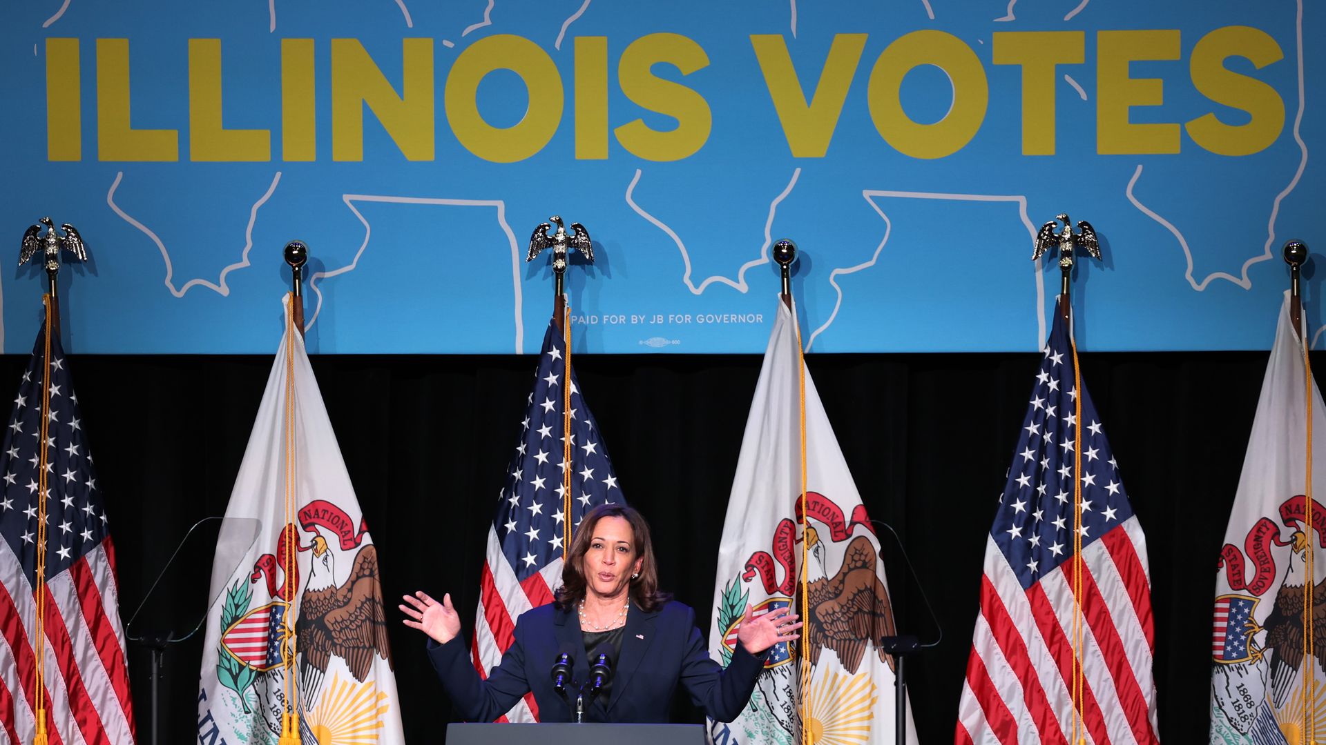 Vice President Harris in a navy blue suit standing behind a podium with the American flag behind her.