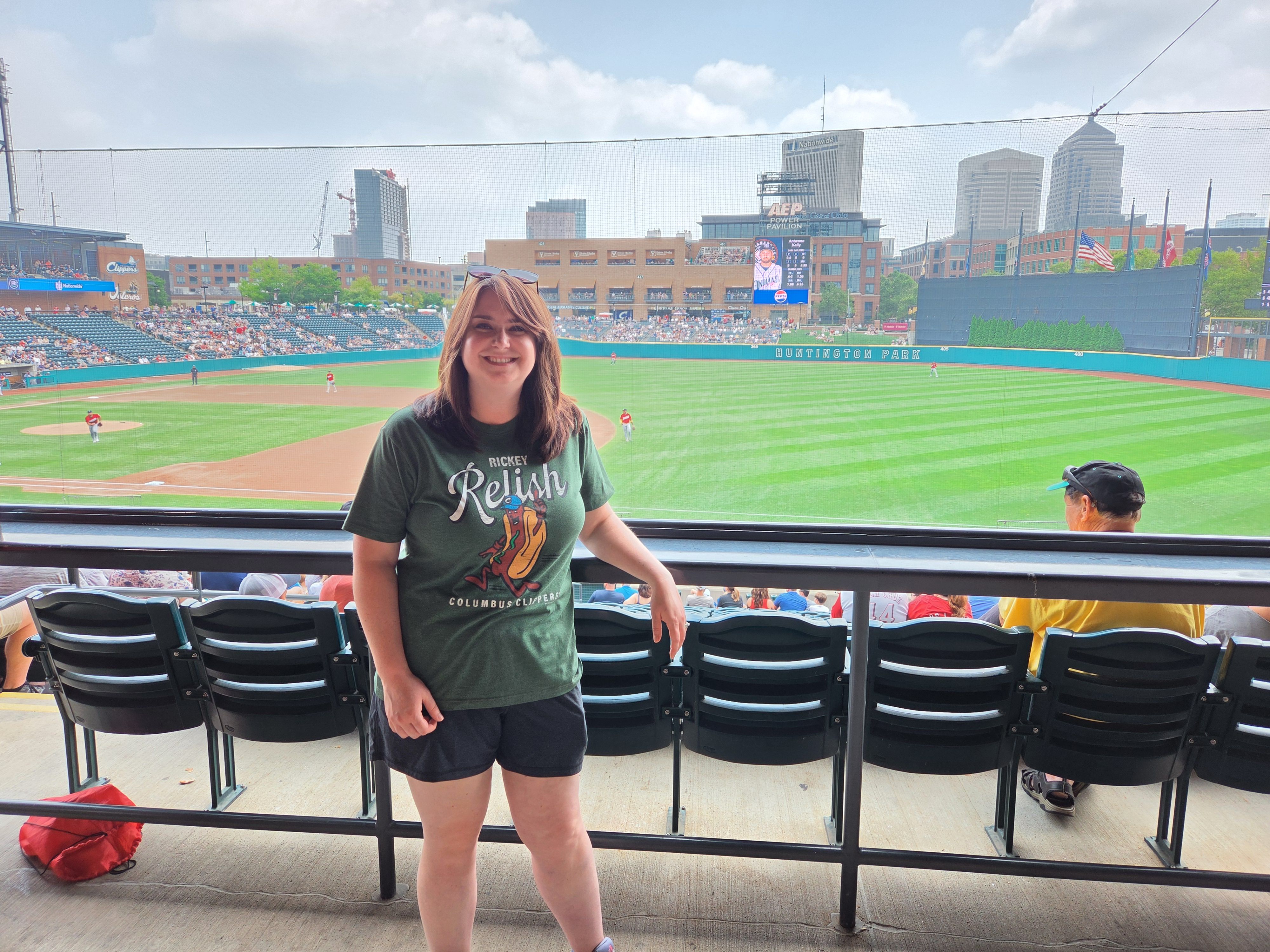 Alissa smiling with sunglasses on her head, wearing a green graphic t-shirt, standing in front of Huntington Park's seating and baseball field, with a scoreboard and city skyline in the background.