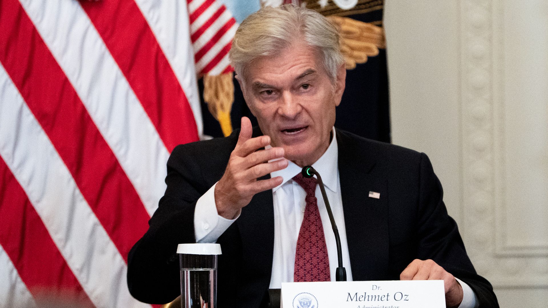 Dr. Mehmet Oz speaking at a podium with a microphone, wearing a dark suit and red tie, with an American flag and presidential seal in the background.