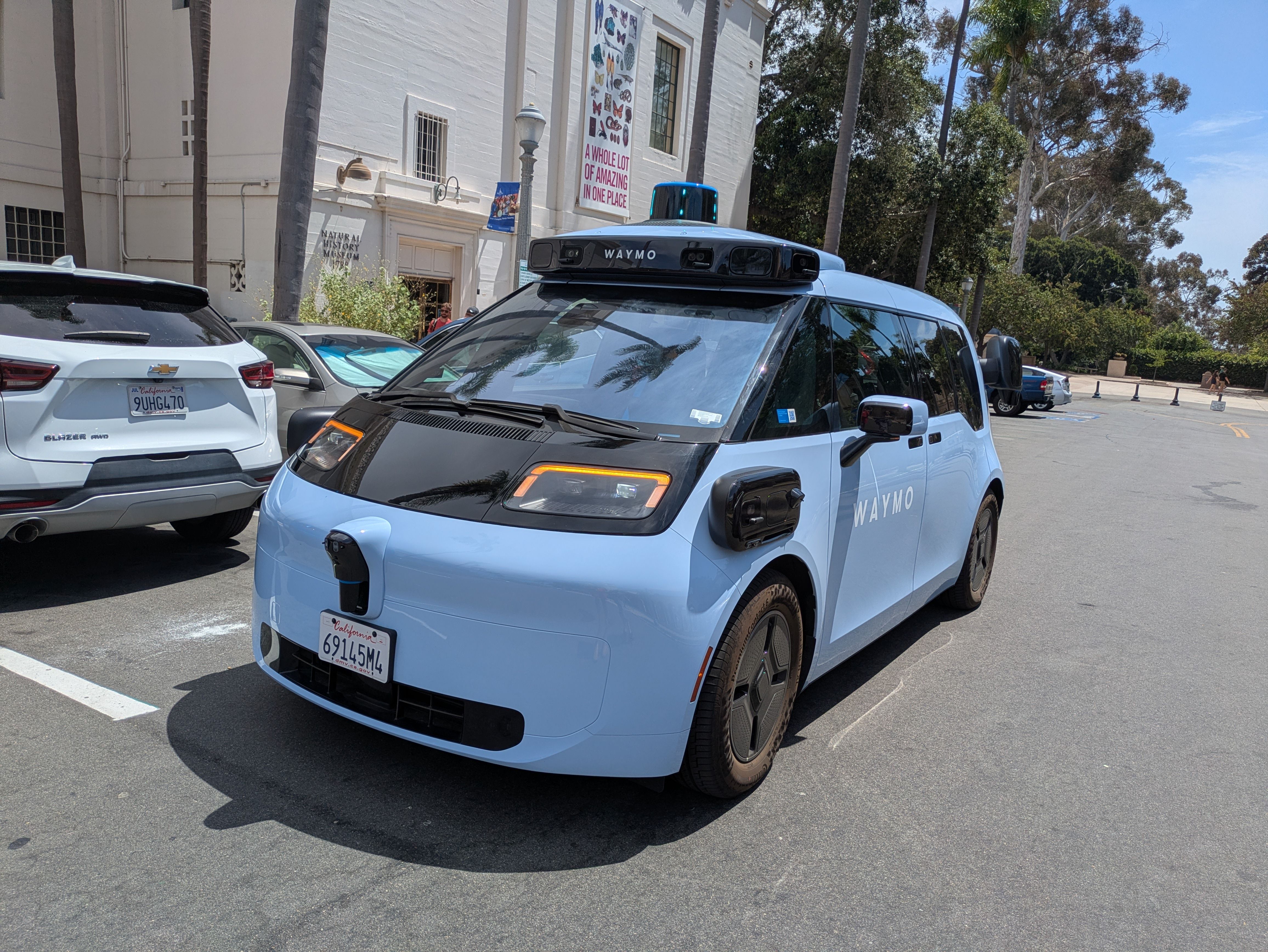 Blue Waymo autonomous van with a blue top light parked in a lot beside a white museum building, flanked by palm trees and other cars.