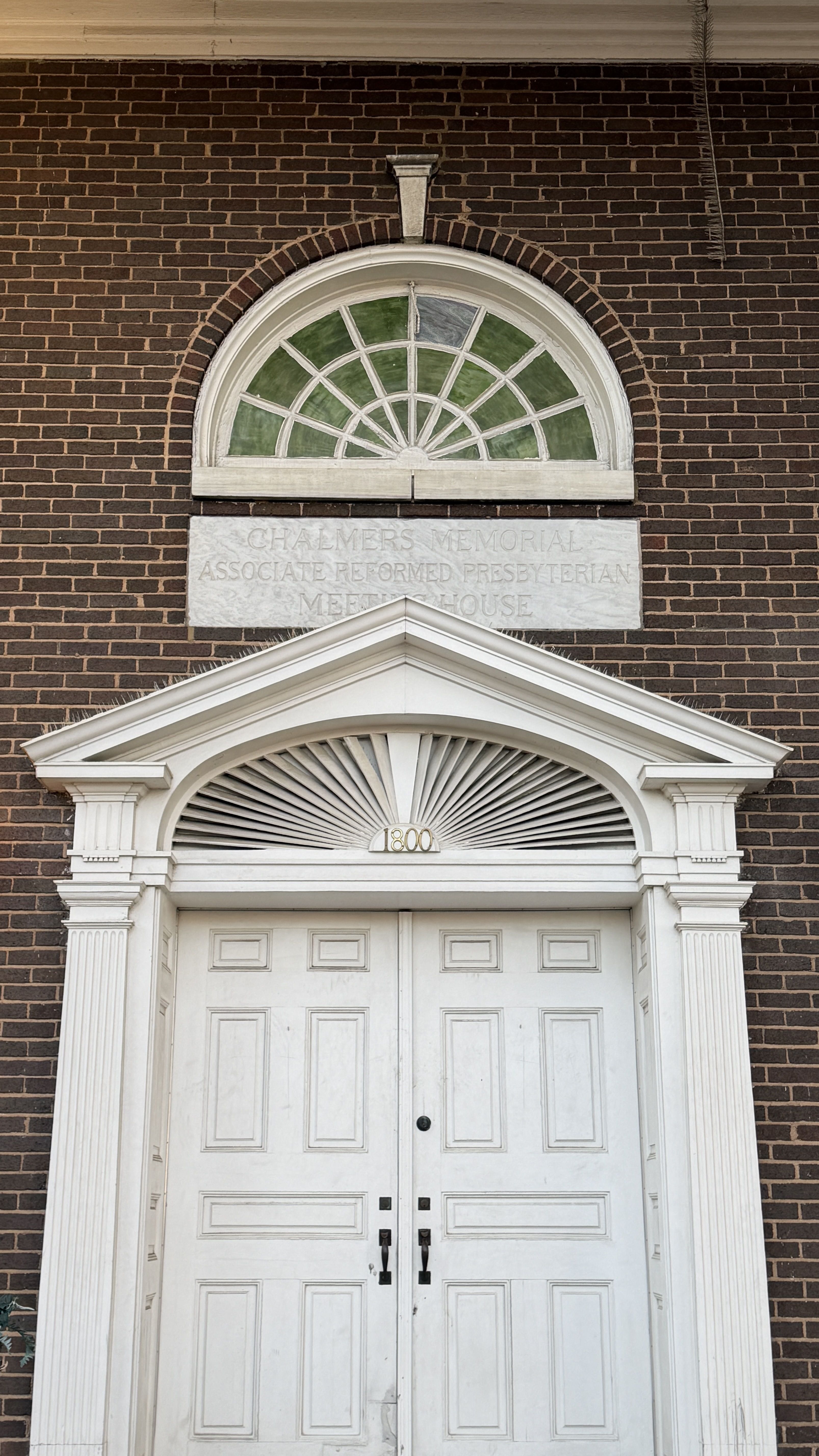 White double doors with decorative molding and '1800' above them, set in a brown brick building with a semicircular window featuring green and white panes above the entry.