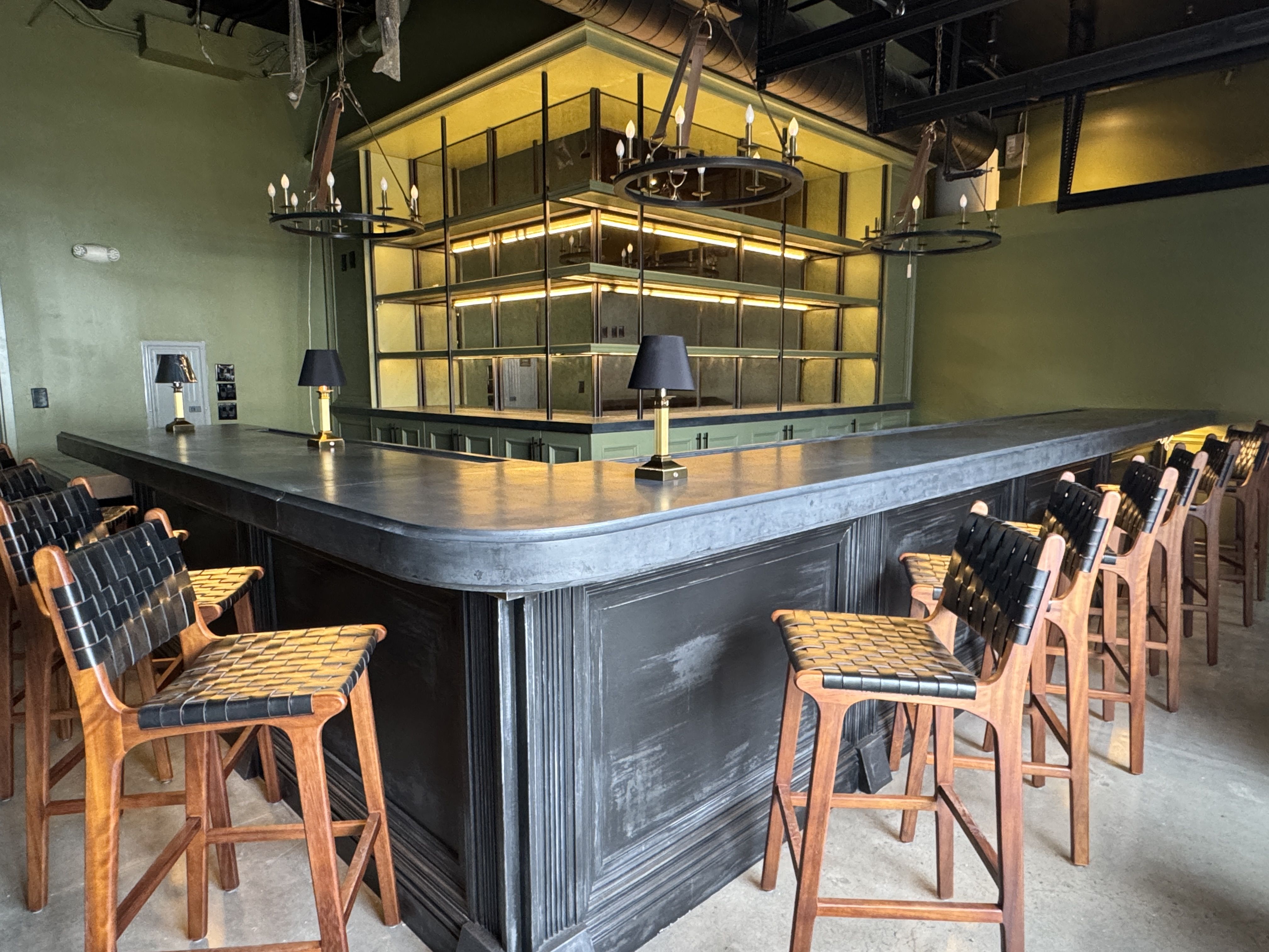 Empty U-shaped bar with black countertop and wooden stools with woven black seats in a green-walled room, illuminated shelves and chandeliers above.