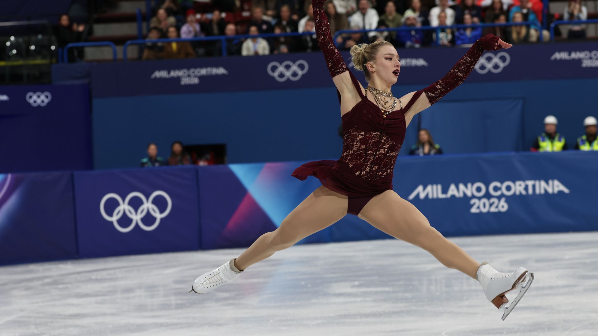 Amber Glenn in a burgundy lace costume jumping with arms raised on ice, with a crowd and 'Milano Cortina 2026' Olympic banners in the background.