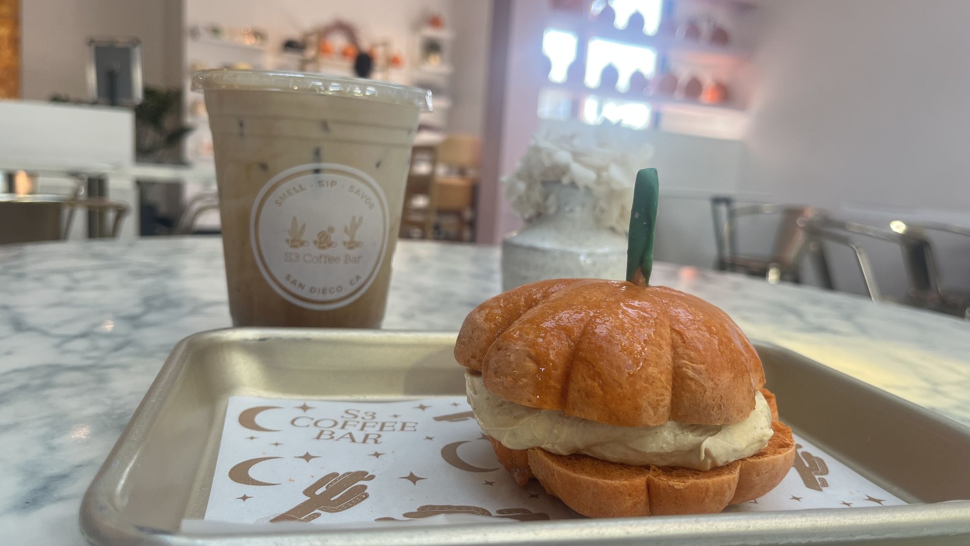 Pumpkin-shaped bagel with cream cheese on a tray with S3 Coffee Bar paper, and an iced coffee in a plastic cup on a marble table inside a cafe with light-colored walls and shelves.