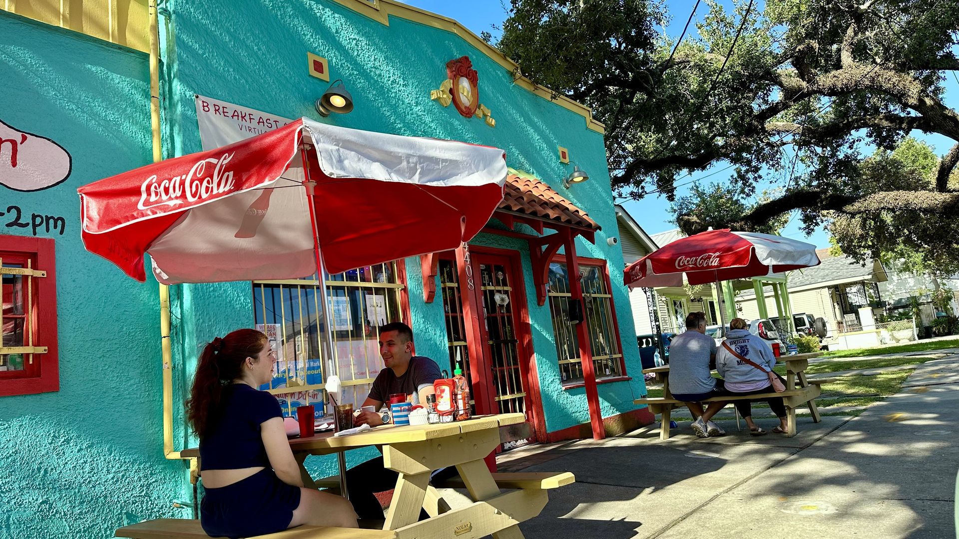 Bright turquoise cafe wall with red trim, red Coca-Cola umbrella, and outdoor wooden tables with people dining. Large tree branches and blue sky frame the sunny street.