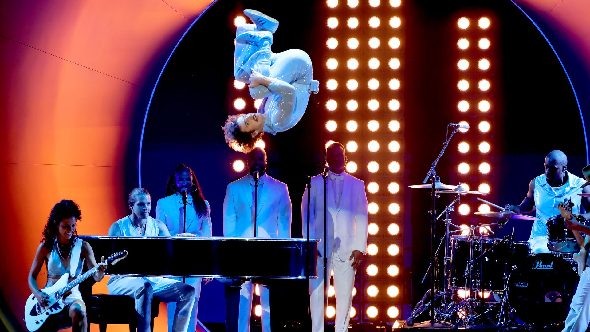 Benson Boone in shiny white suit mid-backflip above a band with guitarist, pianist, three vocalists in white suits, and drummer, all on a stage with bright vertical lights and orange-blue backdrop.