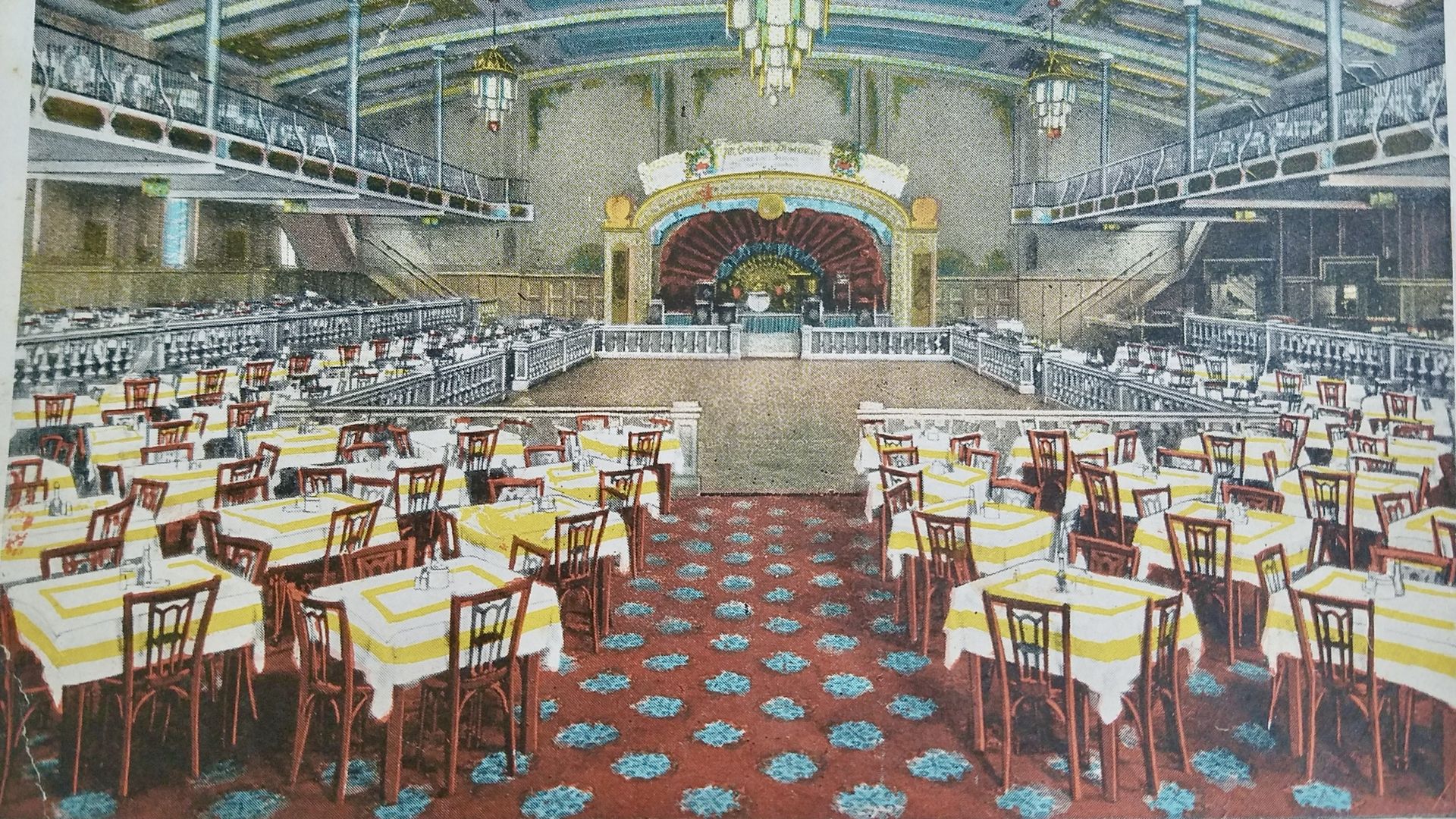 Elegant dining hall with rows of yellow-and-white striped tables and red chairs, a grand ornate stage with gold arches at the far end, and hanging lanterns; balconies run along both sides.