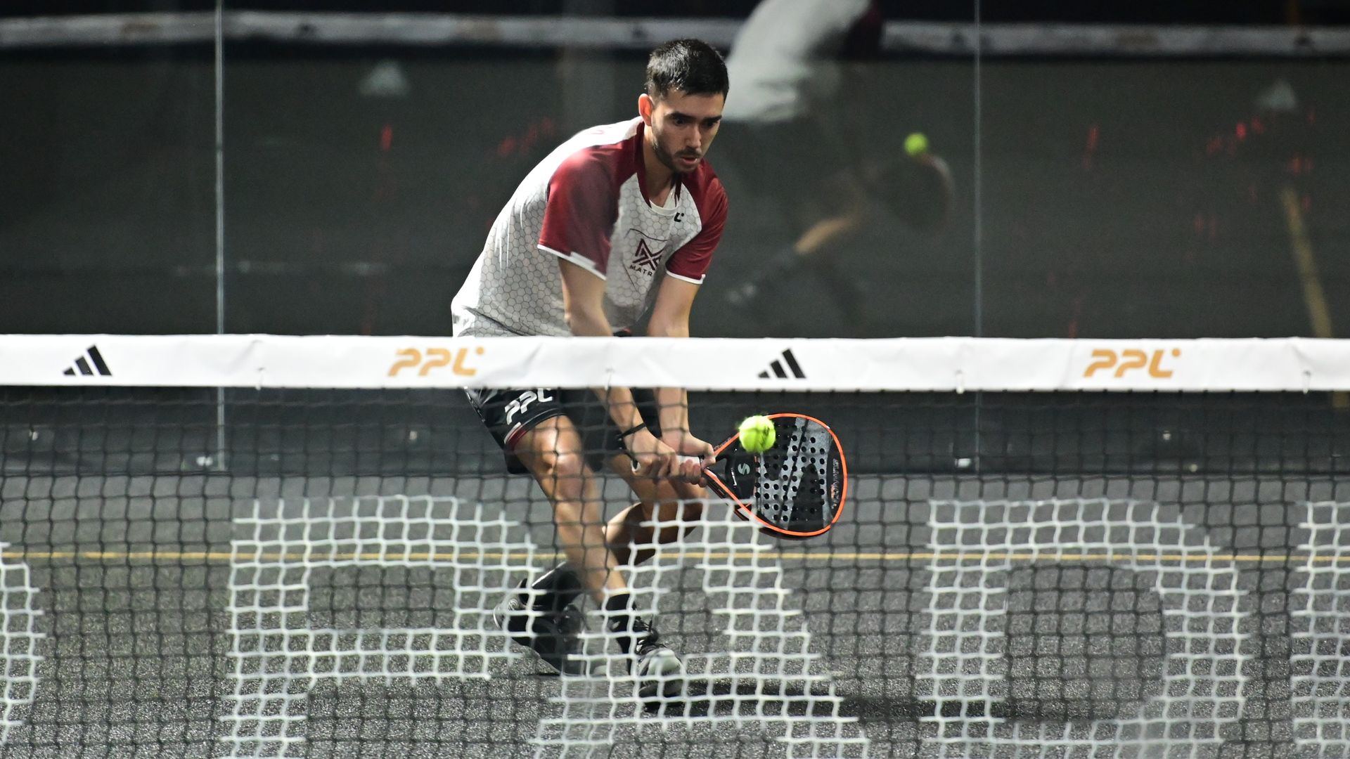 Male squash player in a white and maroon jersey leans forward, hitting a neon yellow ball with an orange racket on an indoor court; glass walls and a white net with PPL logos visible.
