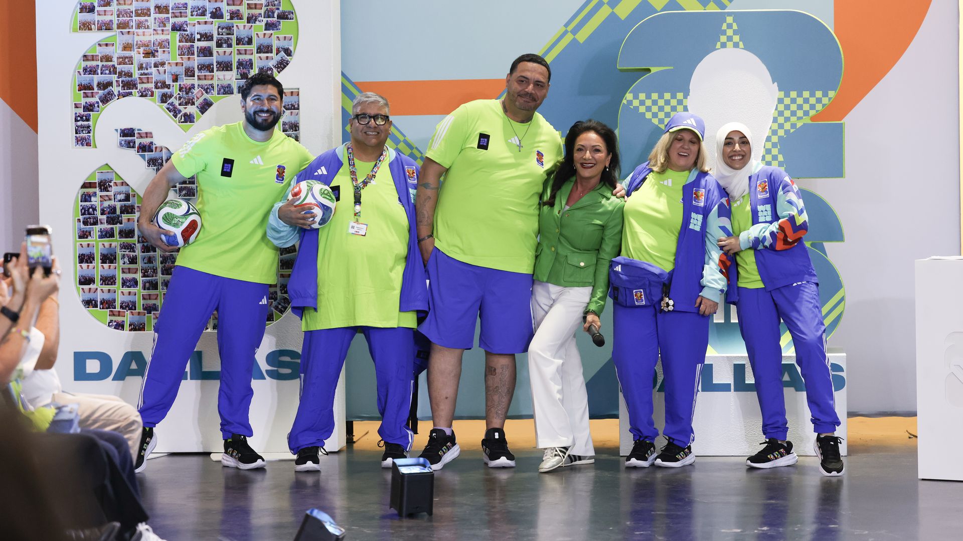 Six people in neon lime-green shirts and blue pants pose on a stage with a large photo collage forming the number 2 and a colorful geometric backdrop; two women wear hijabs; some hold soccer balls.