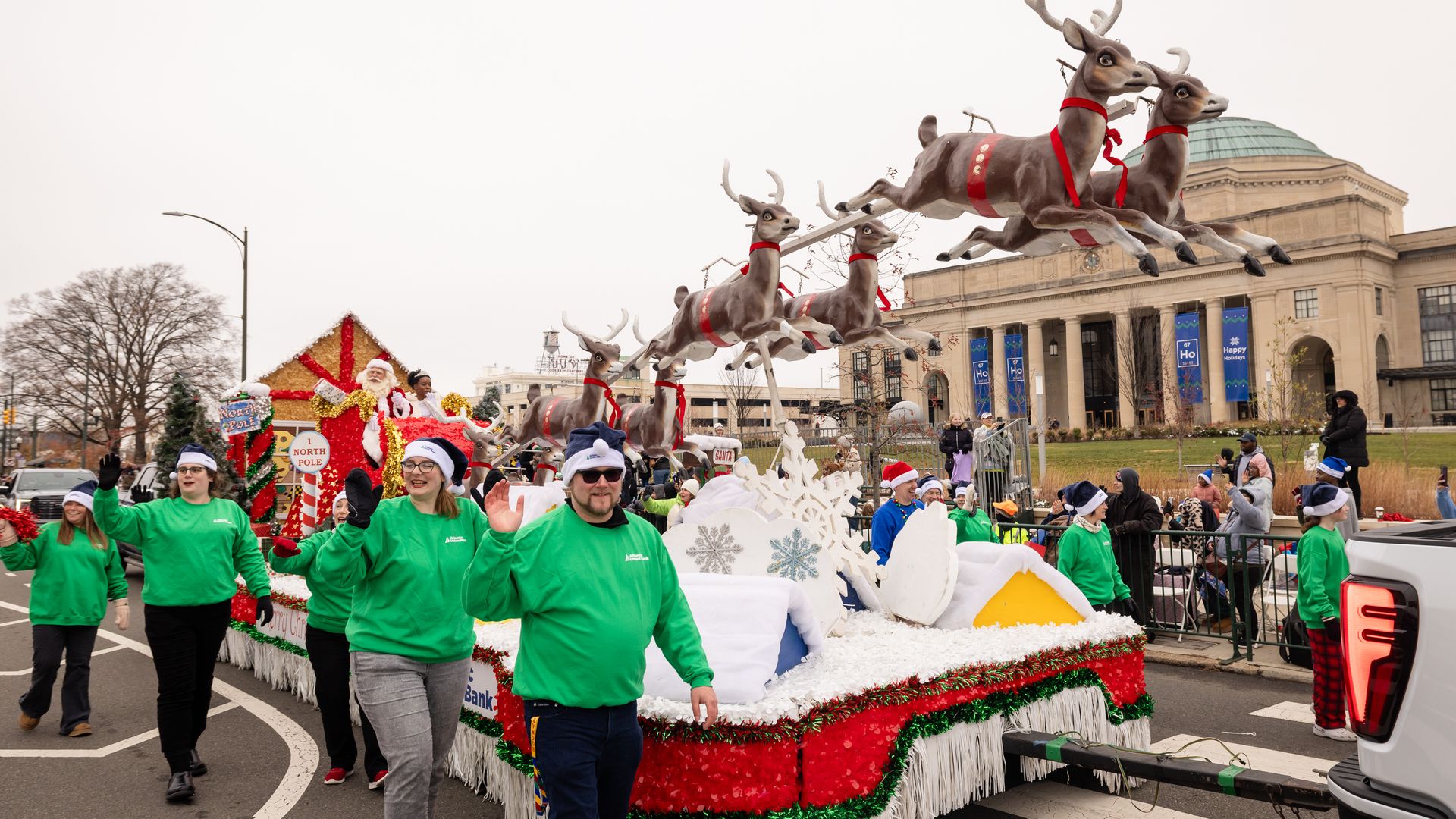 Colorful holiday parade float with jumping reindeer wearing red harnesses, Santa in a wagon, and volunteers in bright green shirts marching along. A domed building rises in the background.
