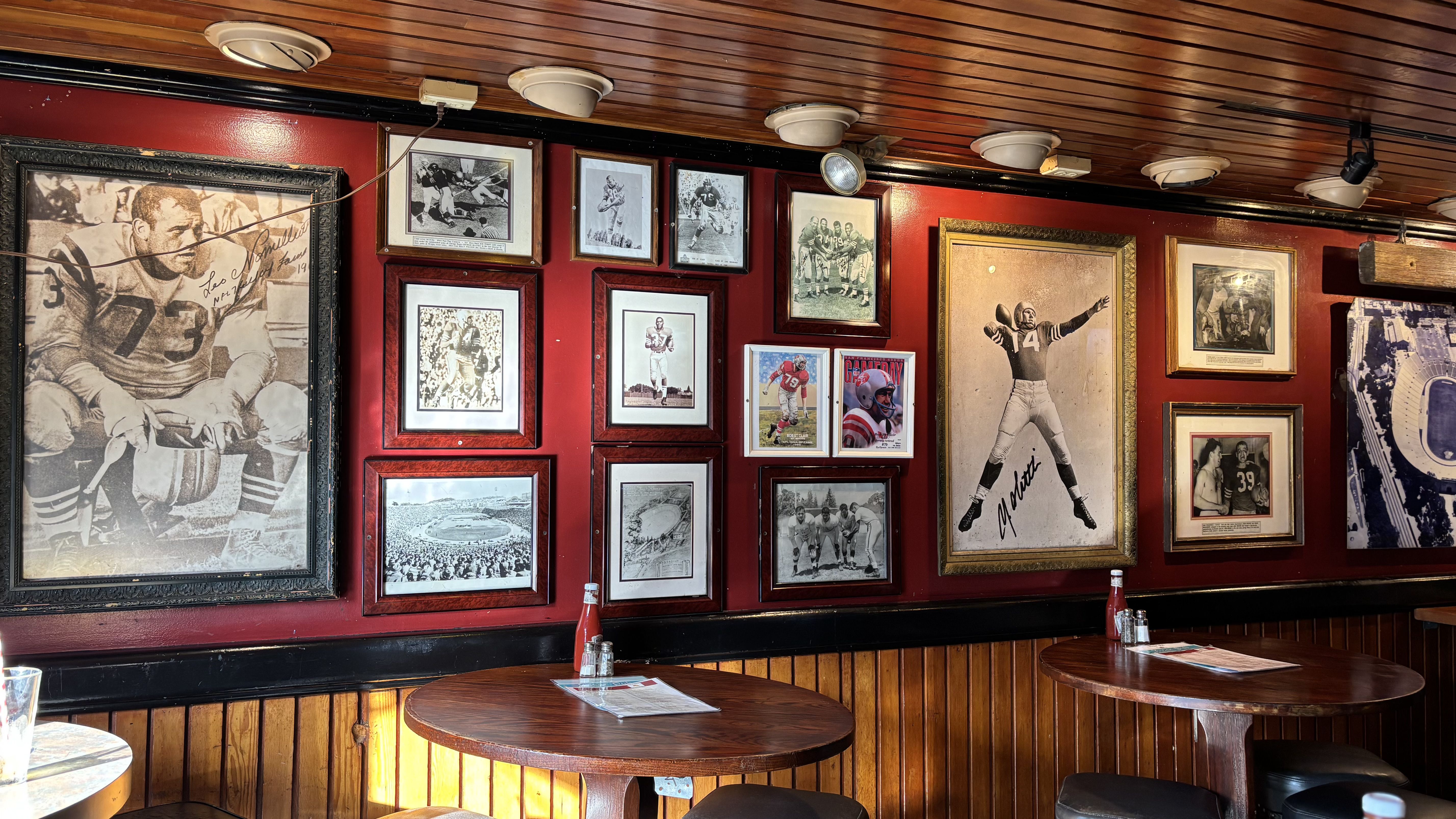 Interior of a sports-themed room with red walls decorated with framed black-and-white vintage football photos and two wooden round tables with menus, ketchup, and salt and pepper.
