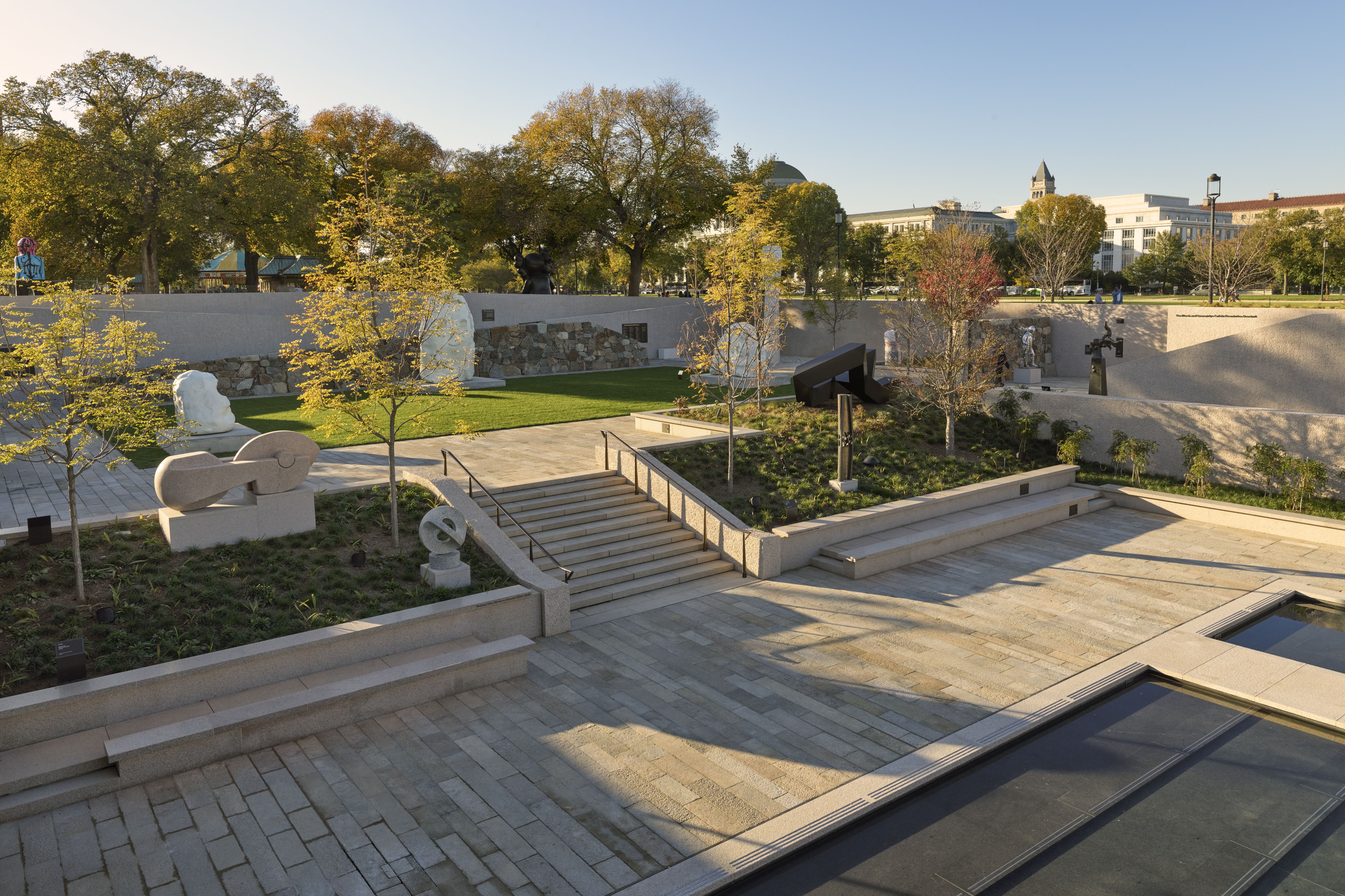 Wide view of an outdoor sculpture park with staircases and beige stone walls, featuring abstract sculptures. Autumn trees frame a modern plaza and a distant classical building beneath a blue sky.