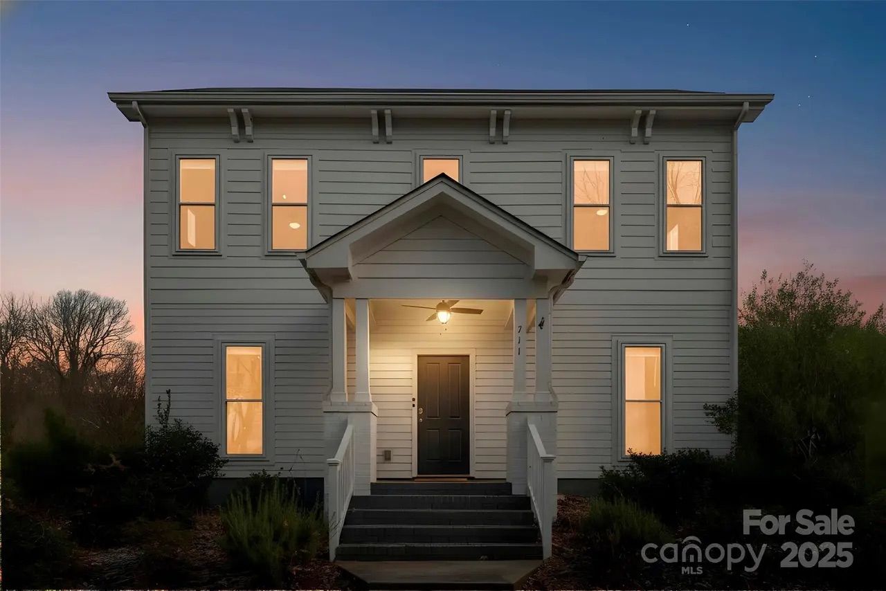 Two-story white house with black door and warm interior lights at dusk, front porch with columns and ceiling fan, surrounded by bushes and trees against a purple-pink sky.