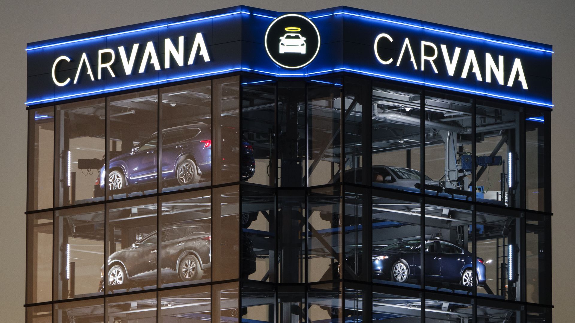 Nighttime view of a glass-front Carvana building with blue neon trim and large CARVANA signs; a circular logo featuring a car with a halo sits above, while cars are visible on multiple floors inside.