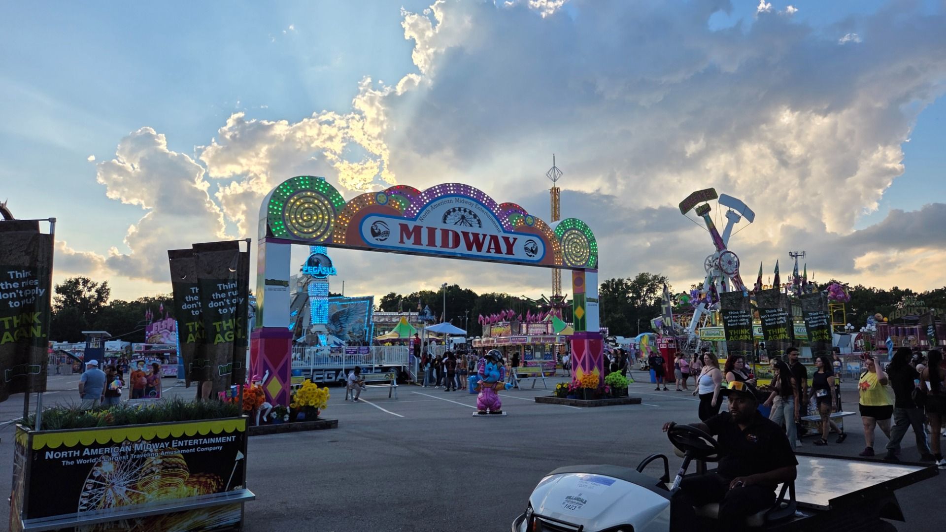 Colorful carnival entrance under a blue, partly cloudy sky. A large lit MIDWAY arch with spiraling LEDs, rides to the right, banners and stalls, and people strolling through the fairgrounds.