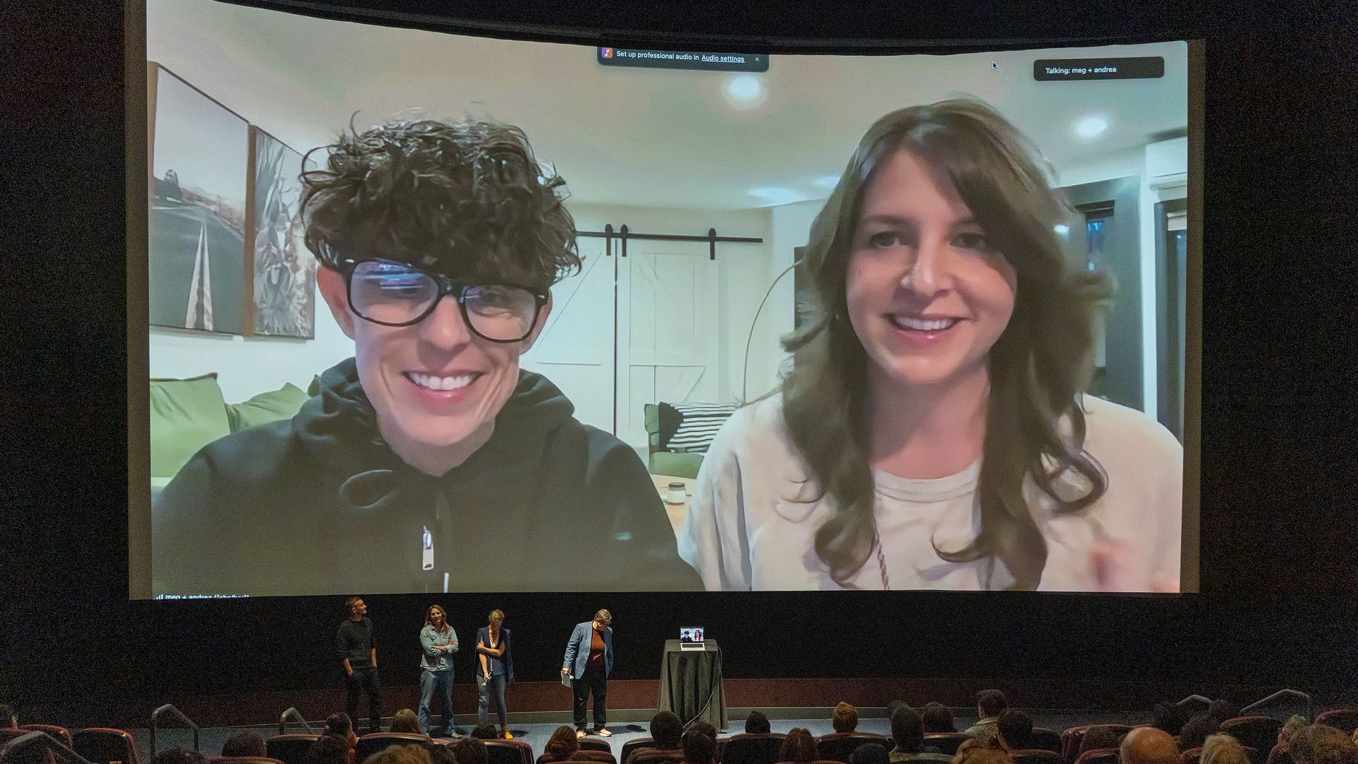 Dark theater with a giant screen showing a video call: Andrea Gibson and Megan Falley. A small group stands on stage below, audience seated.