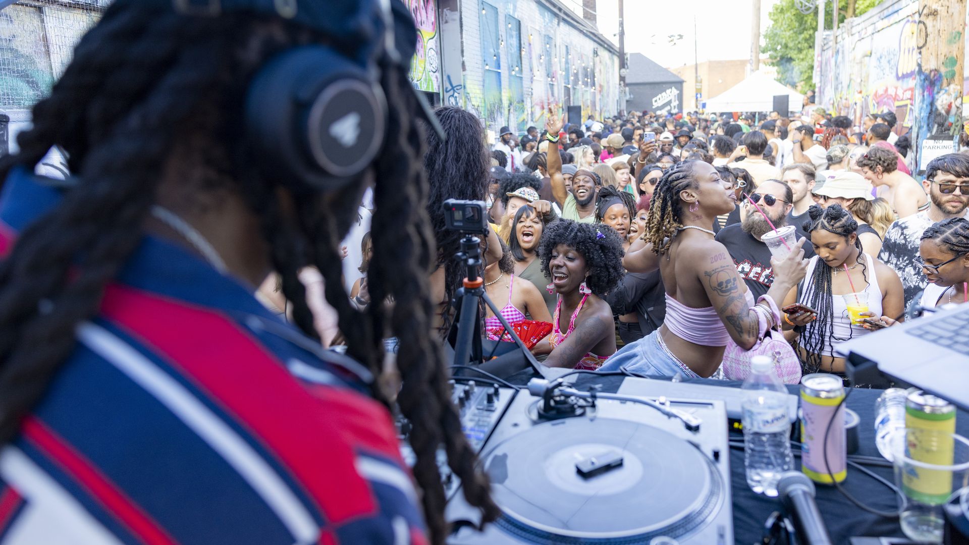 DJ with headphones in red and blue shirt playing music on turntables at an outdoor street party with a diverse crowd dancing and enjoying drinks near graffiti-covered walls.