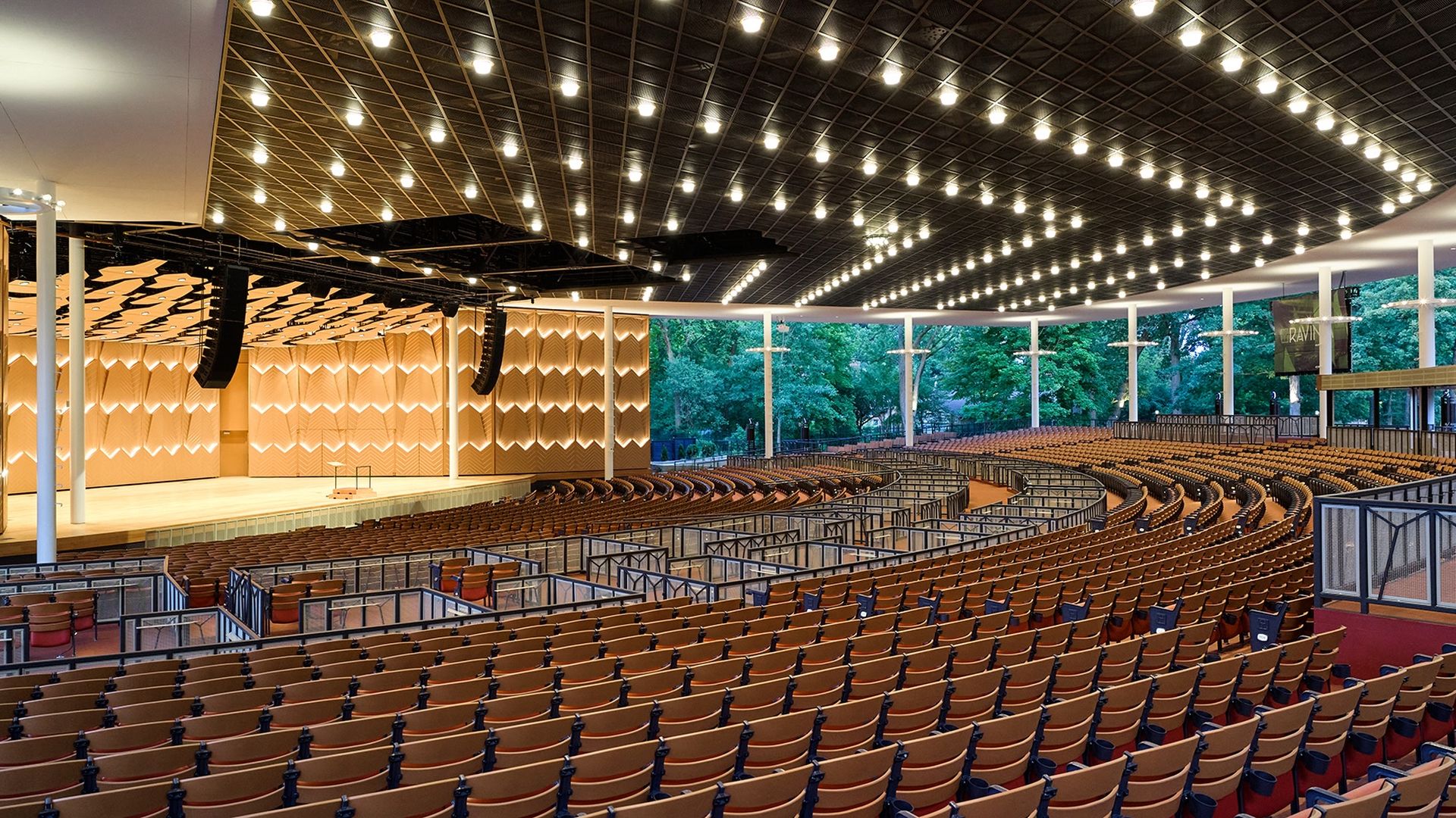 Wide view of a modern concert hall with curved rows of brown seats, a wood-patterned stage wall, white columns, and a grid of bright ceiling lights. Large windows reveal green trees outside.