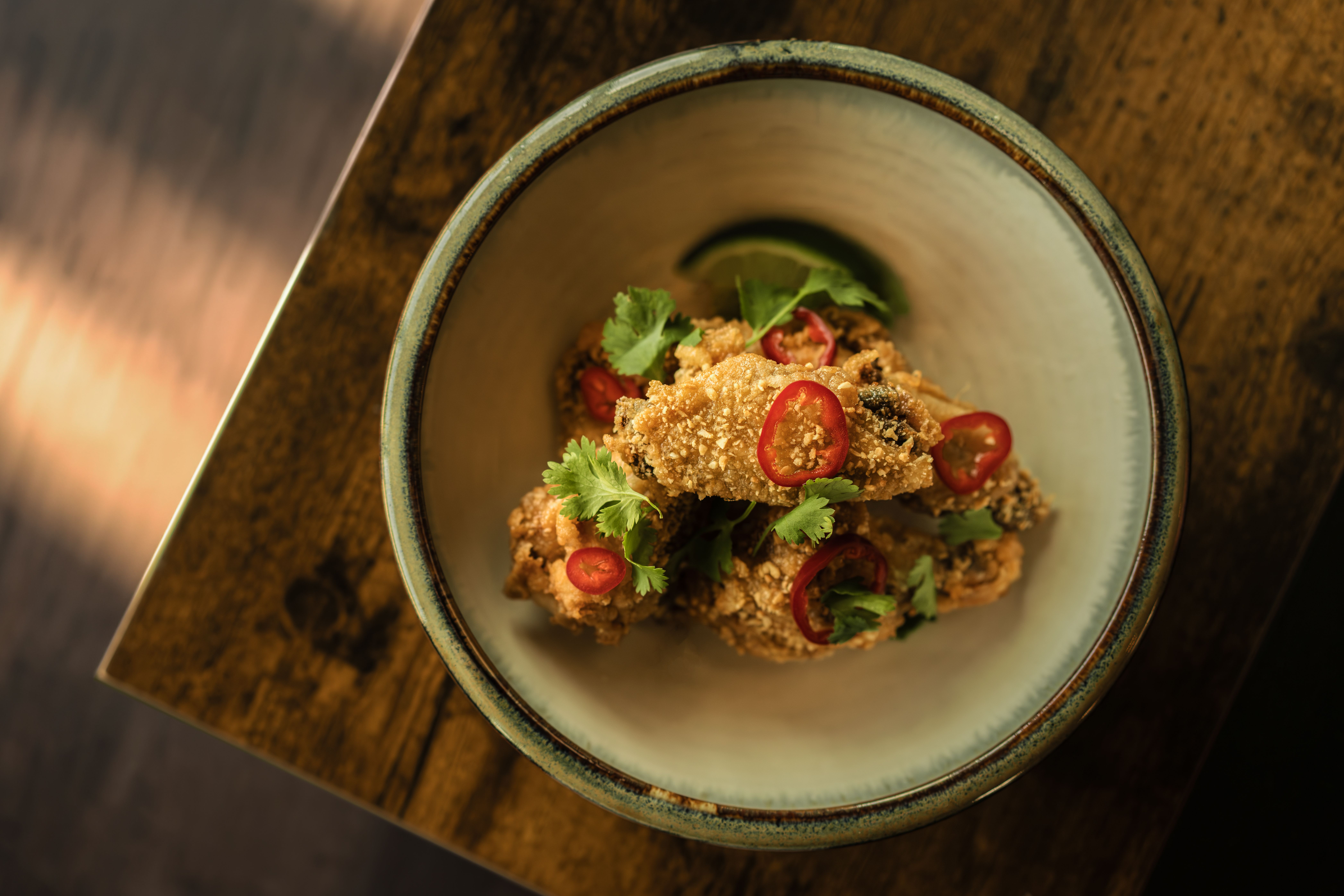 Overhead view of a ceramic bowl on a dark wooden table, filled with crispy fried chicken bites, garnished with red chili rings and fresh cilantro.